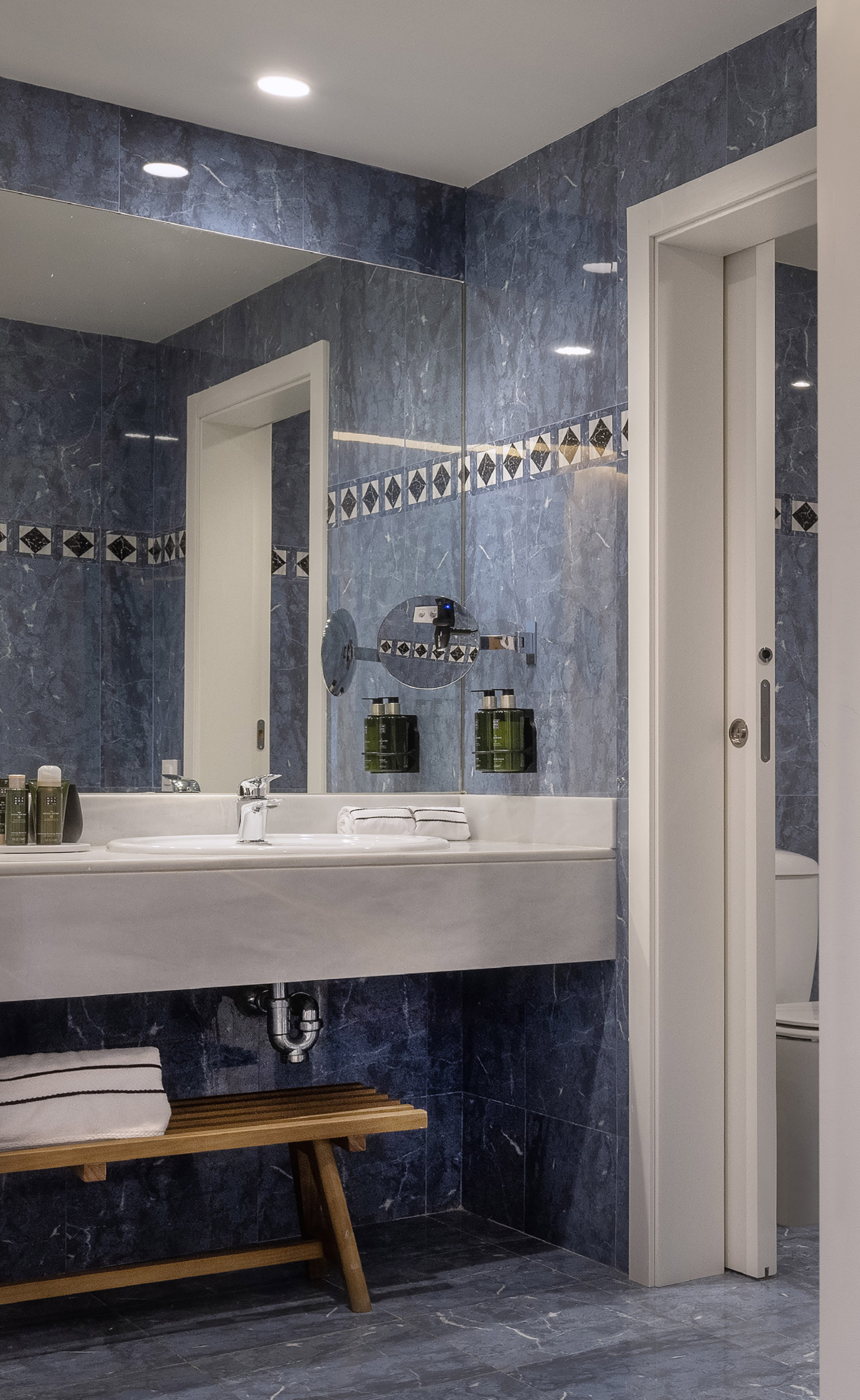 Modern blue marble bathroom with white vanity, large mirror, and wood bench.