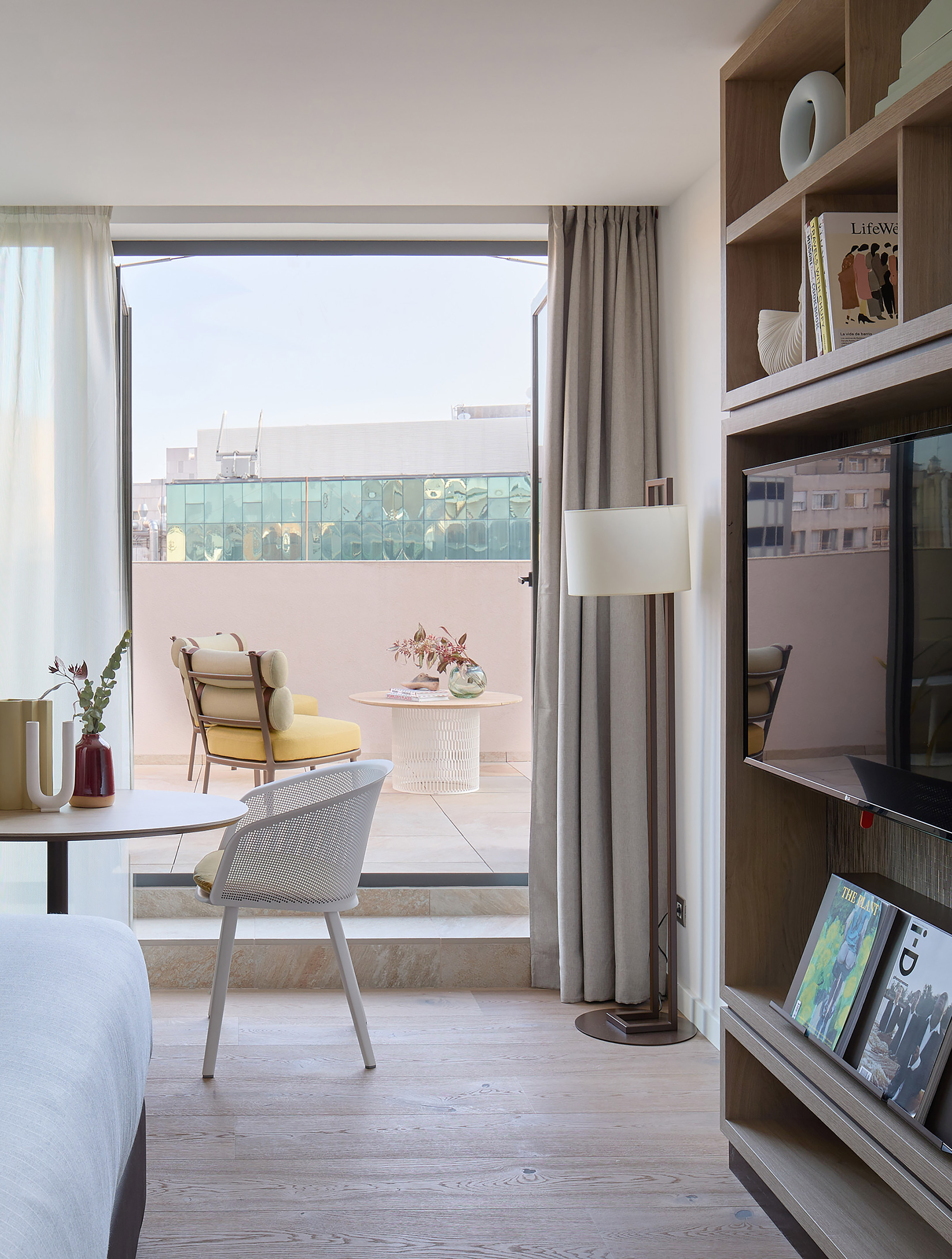 Modern hotel room with balcony, outdoor seating, built-in TV, and wood floor.