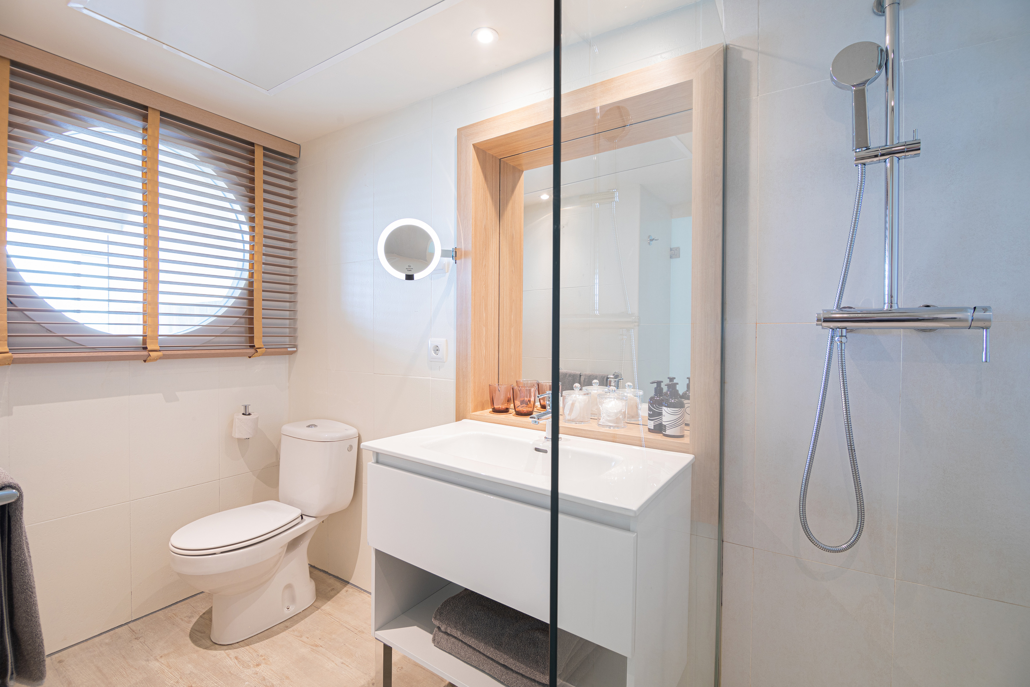 Modern hotel bathroom with white vanity, toilet, shower, and window blinds.