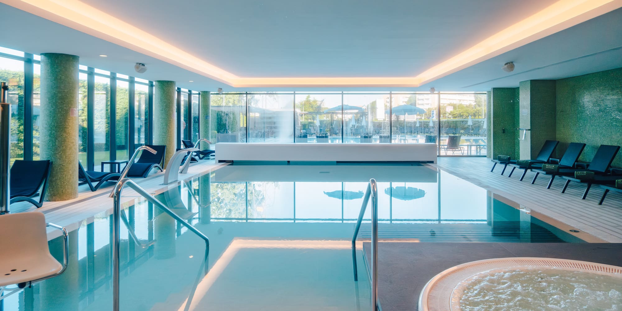 Luxury indoor spa pool with hot tub, lounge chairs, and large windows.