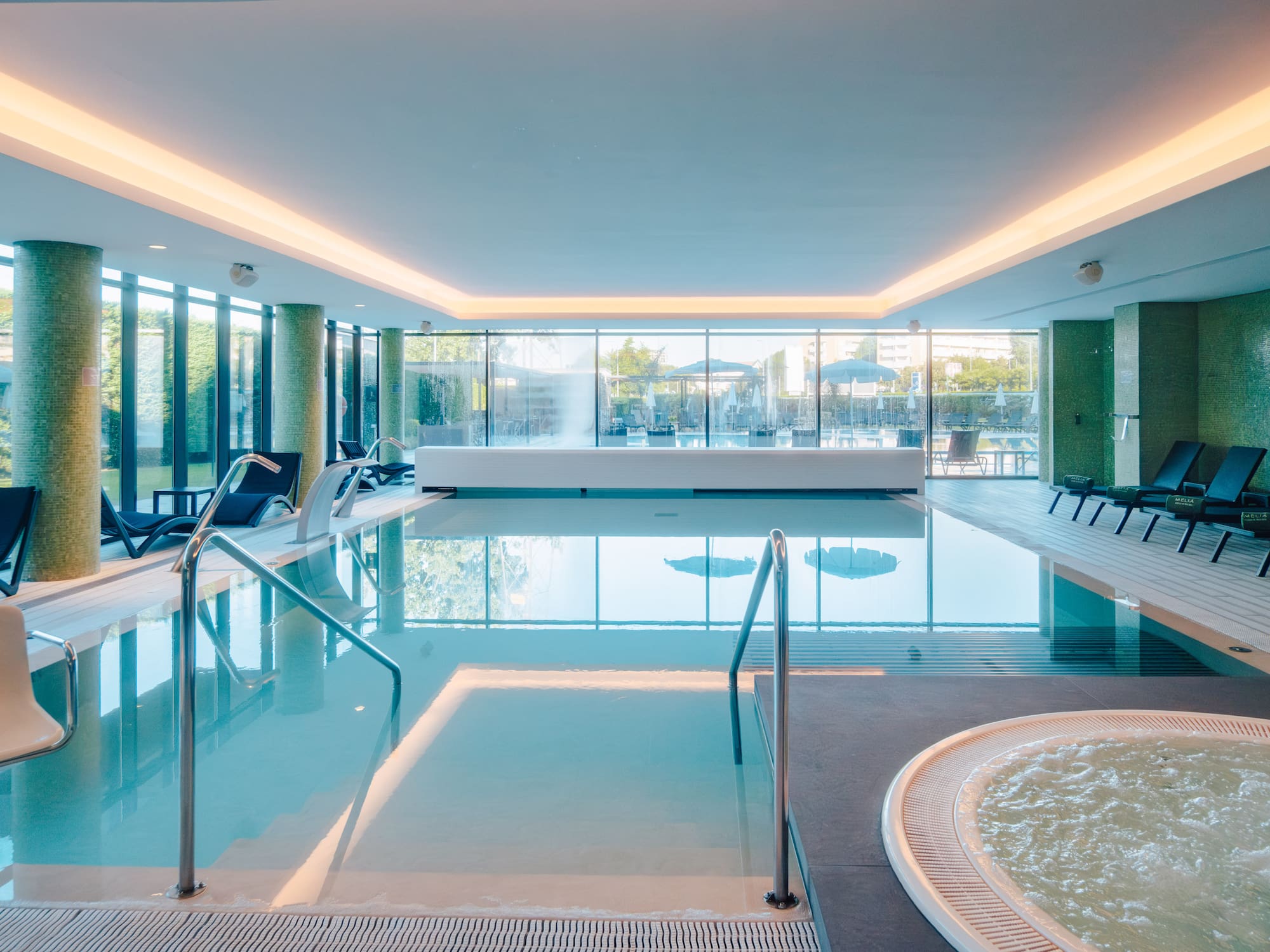 Modern indoor hotel pool with lounge seating, jacuzzi, and glass walls.