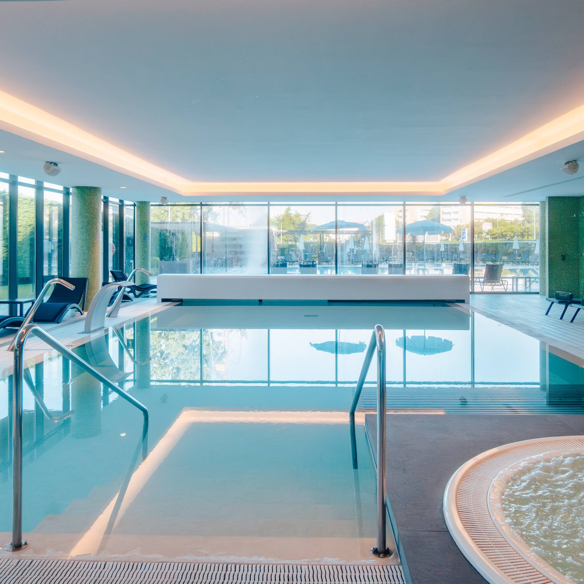 Bright indoor spa pool with hot tub, lounge chairs, and large windows.