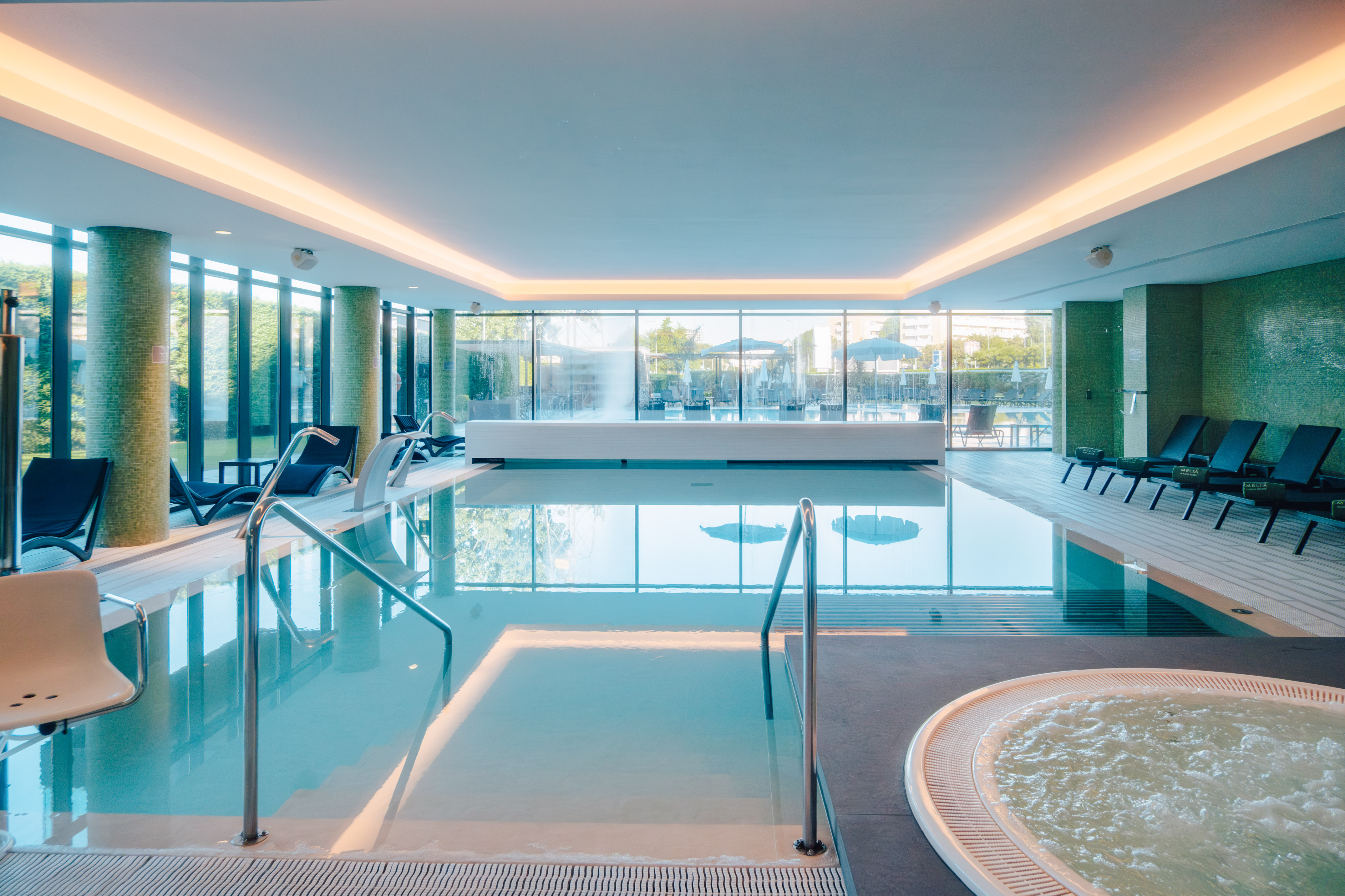 Modern indoor hotel pool with lounge seating, jacuzzi, and glass walls.