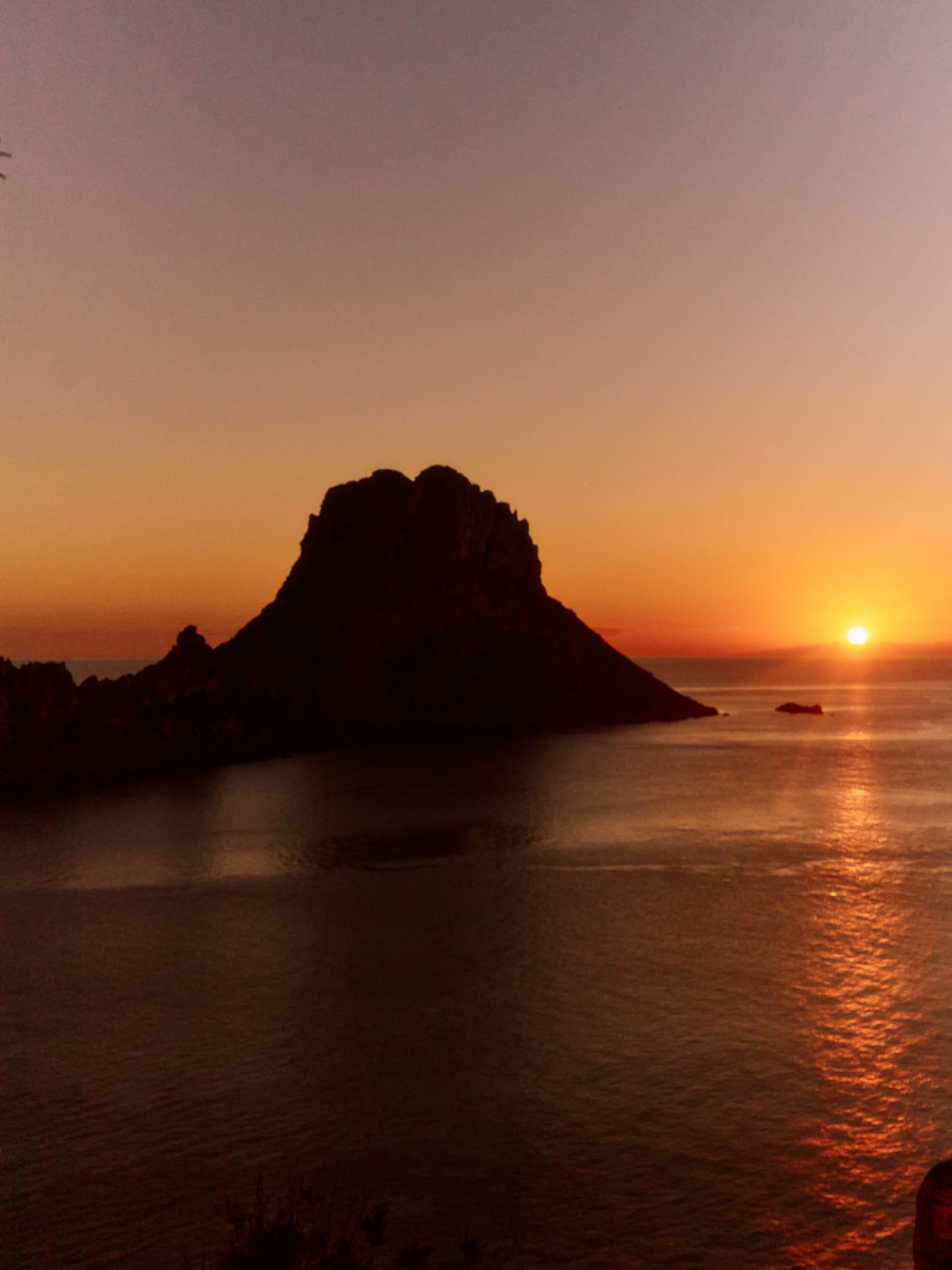 Sunset over ocean with dark rocky island silhouette, reflecting orange sky.