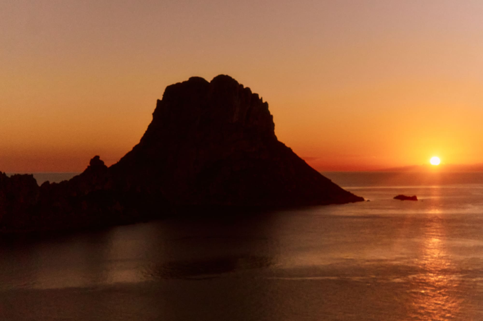 Sunset over ocean with dark rocky island silhouette, reflecting orange sky.