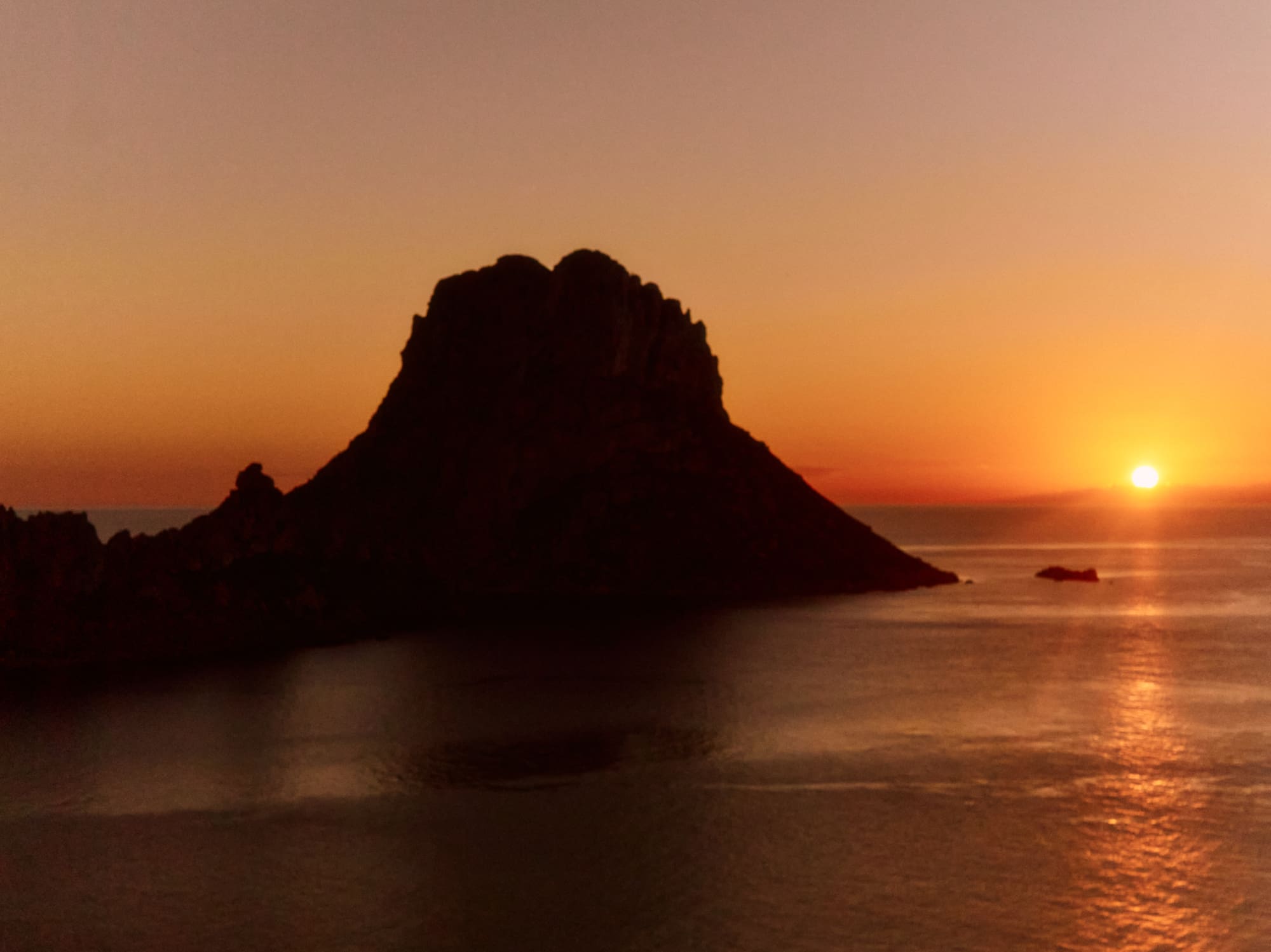 Sunset over ocean with dark rocky island silhouette, reflecting orange sky.
