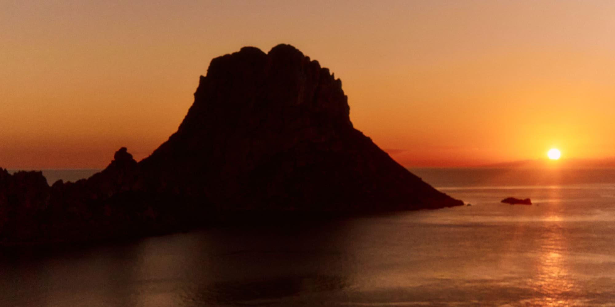 Sunset over ocean with dark rocky island silhouette, reflecting orange sky.