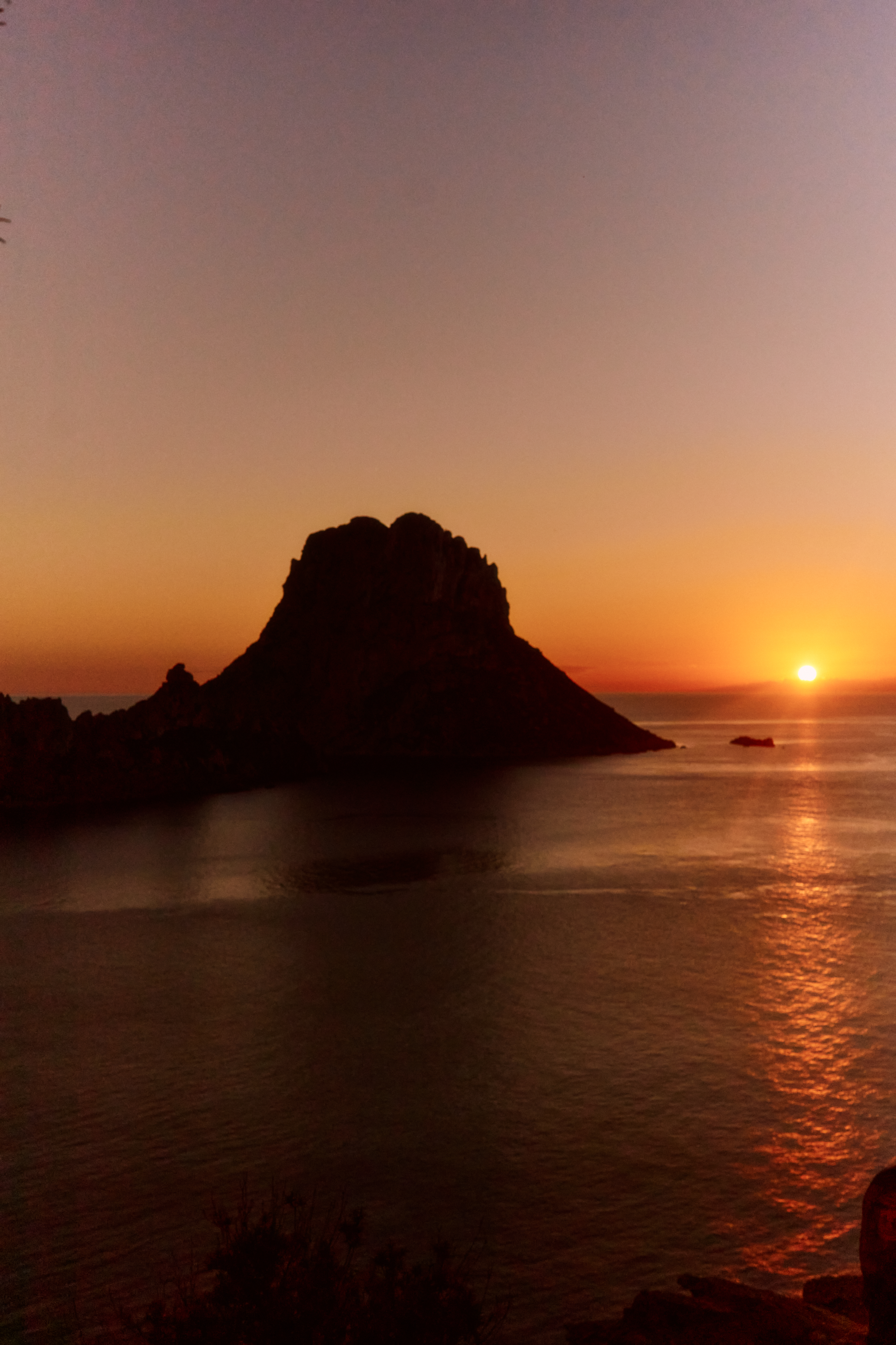 Sunset over ocean with dark rocky island silhouette, reflecting orange sky.