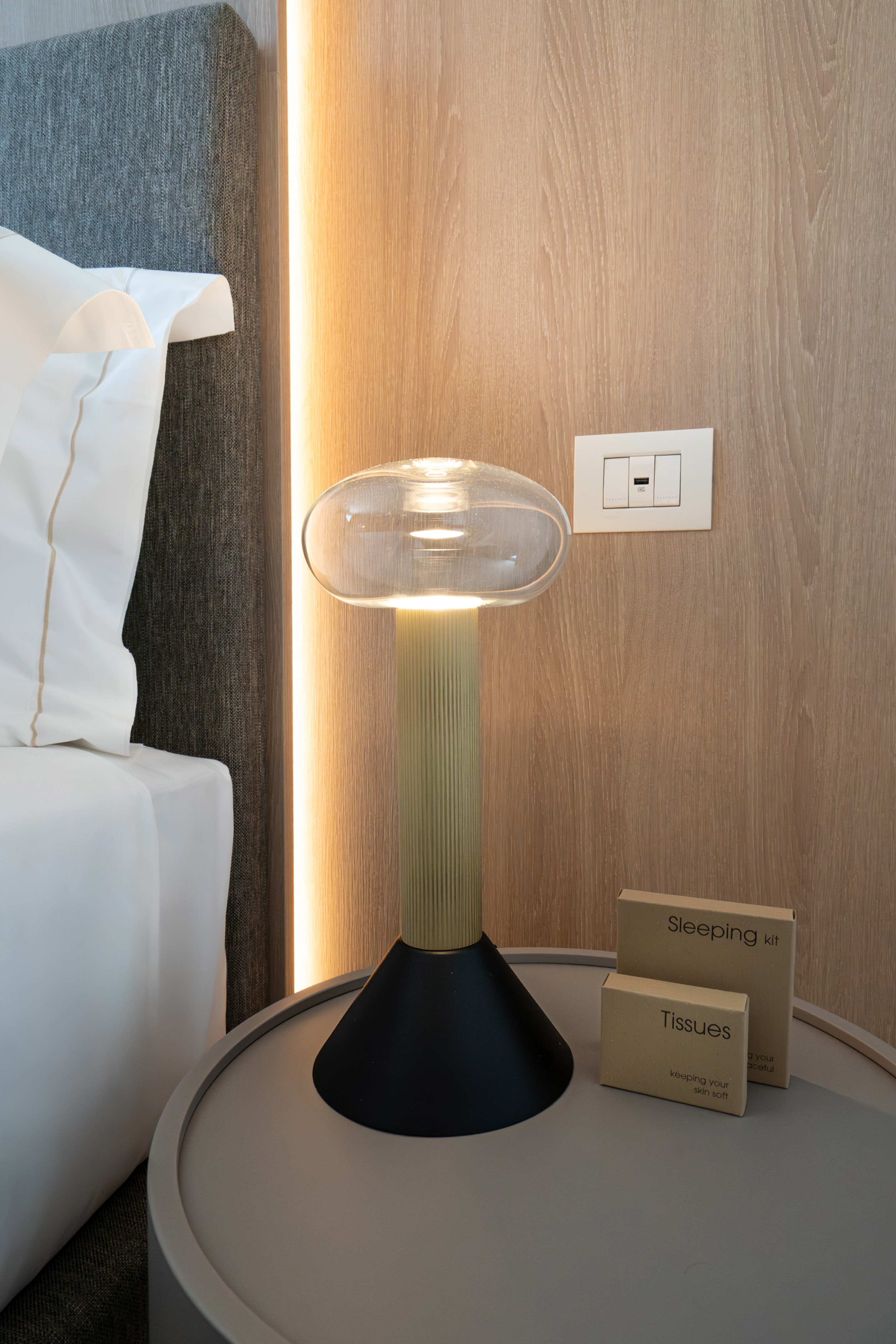 Modern hotel bedside table with contemporary lamp, sleeping kit, and tissues.