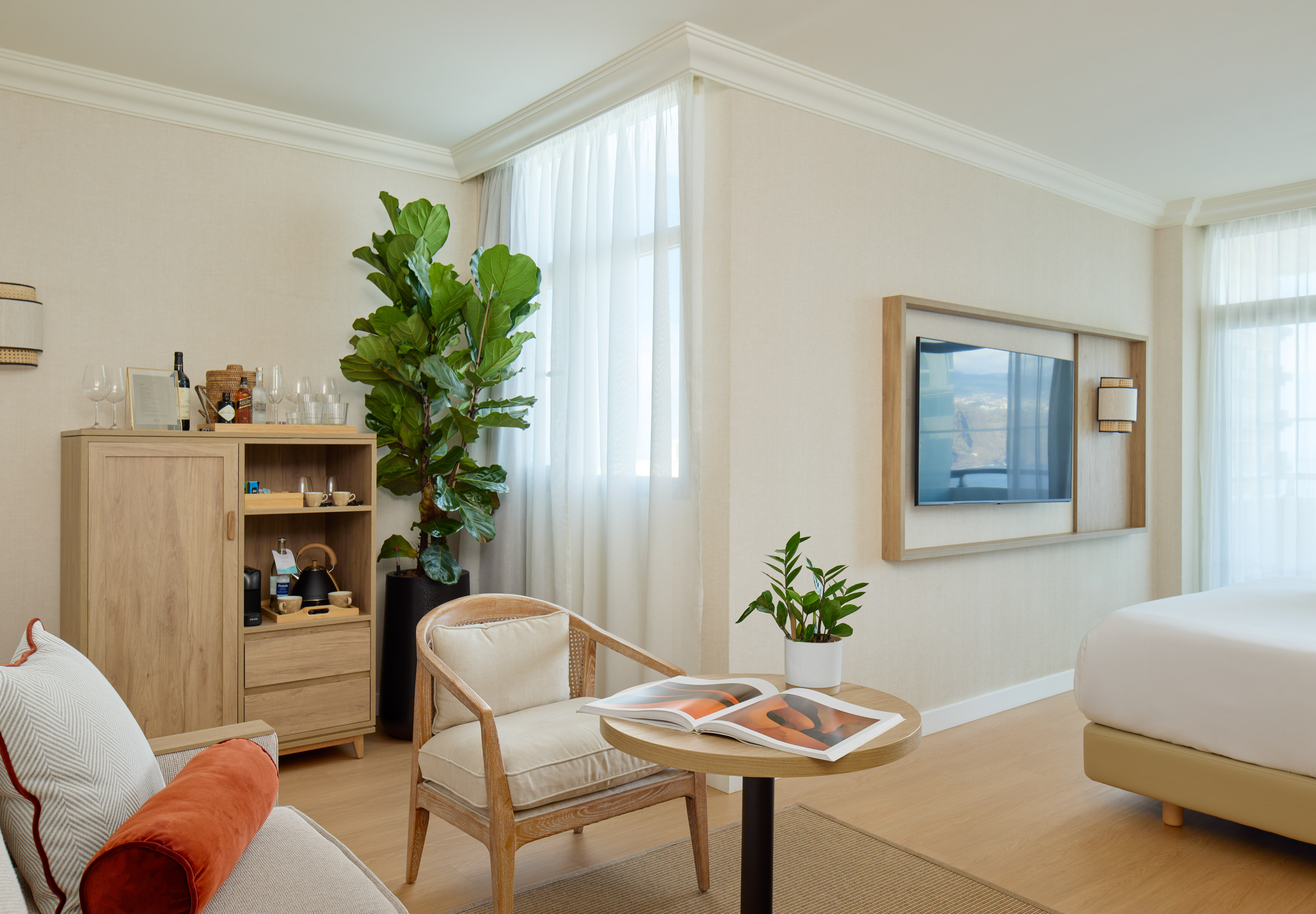 Modern hotel suite with neutral decor, seating area, TV, and large window.
