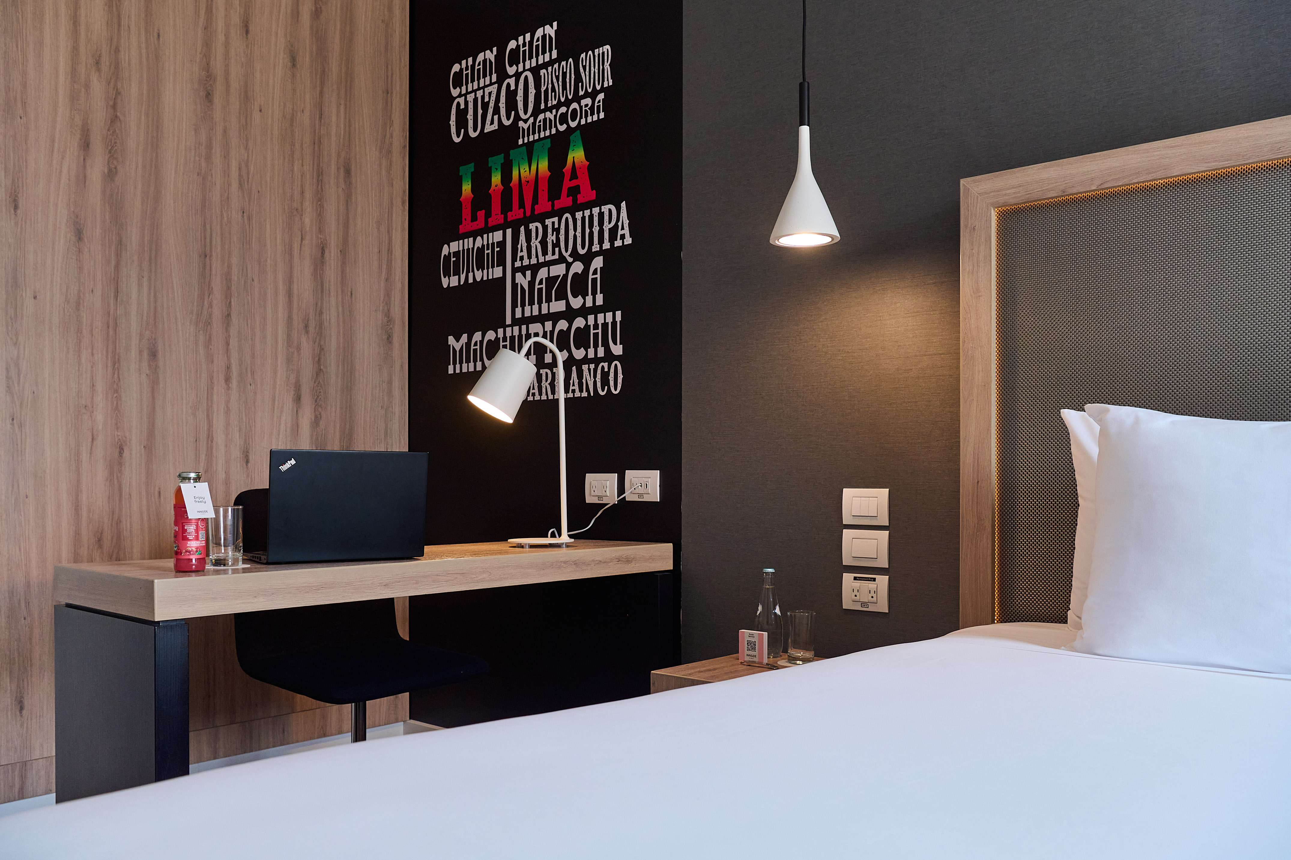 Modern hotel room: bed, wooden desk with laptop, Peruvian city name wall art.