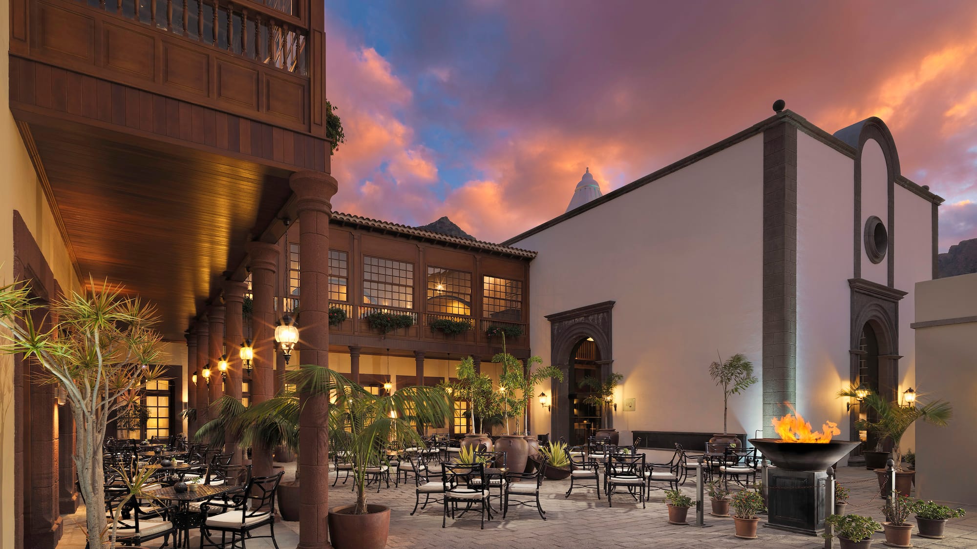 Resort courtyard with fire pit, outdoor dining tables, and colorful sunset sky.