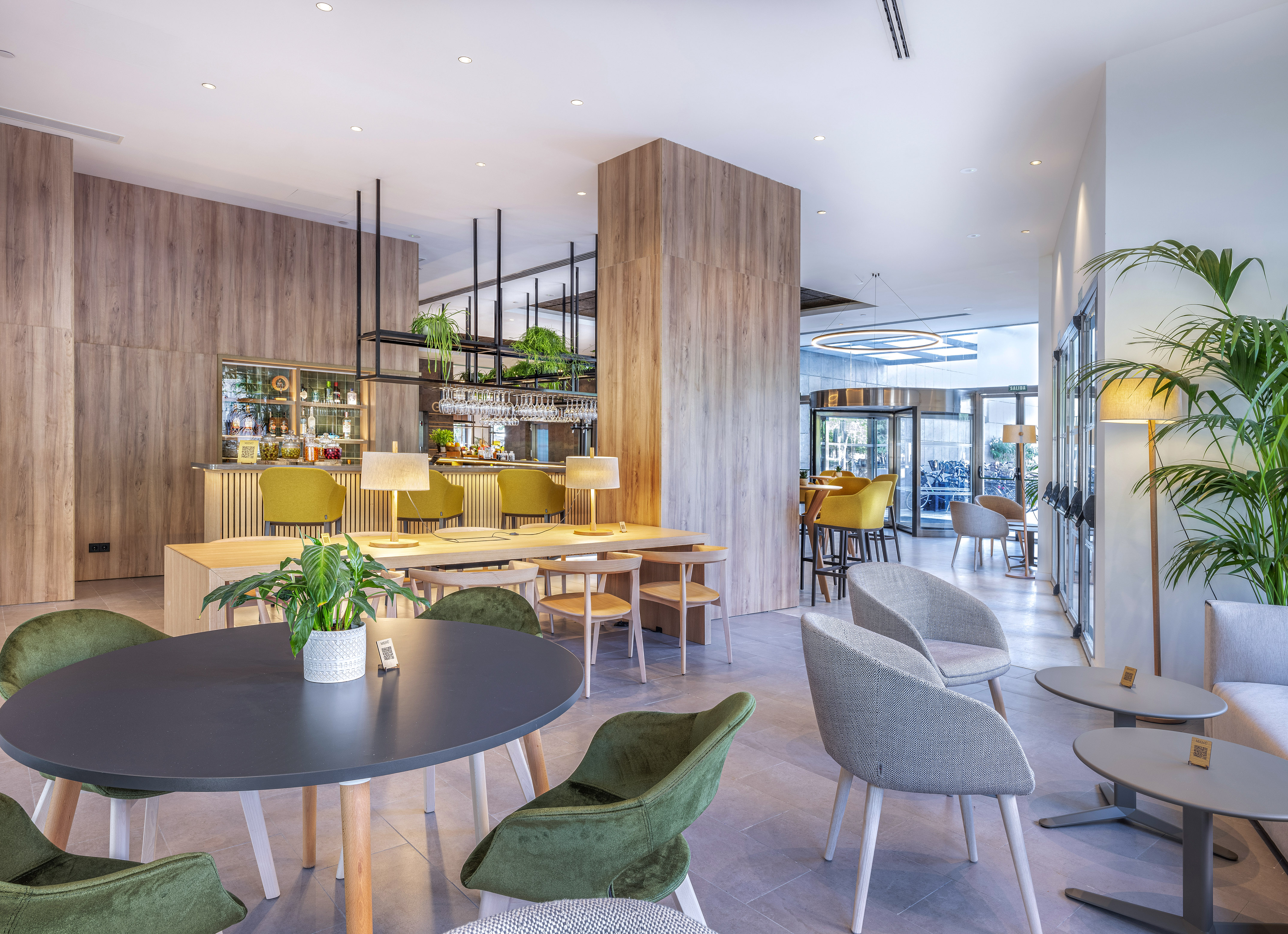 Contemporary hotel bar & lounge with wood panels, green and grey seating, plants.