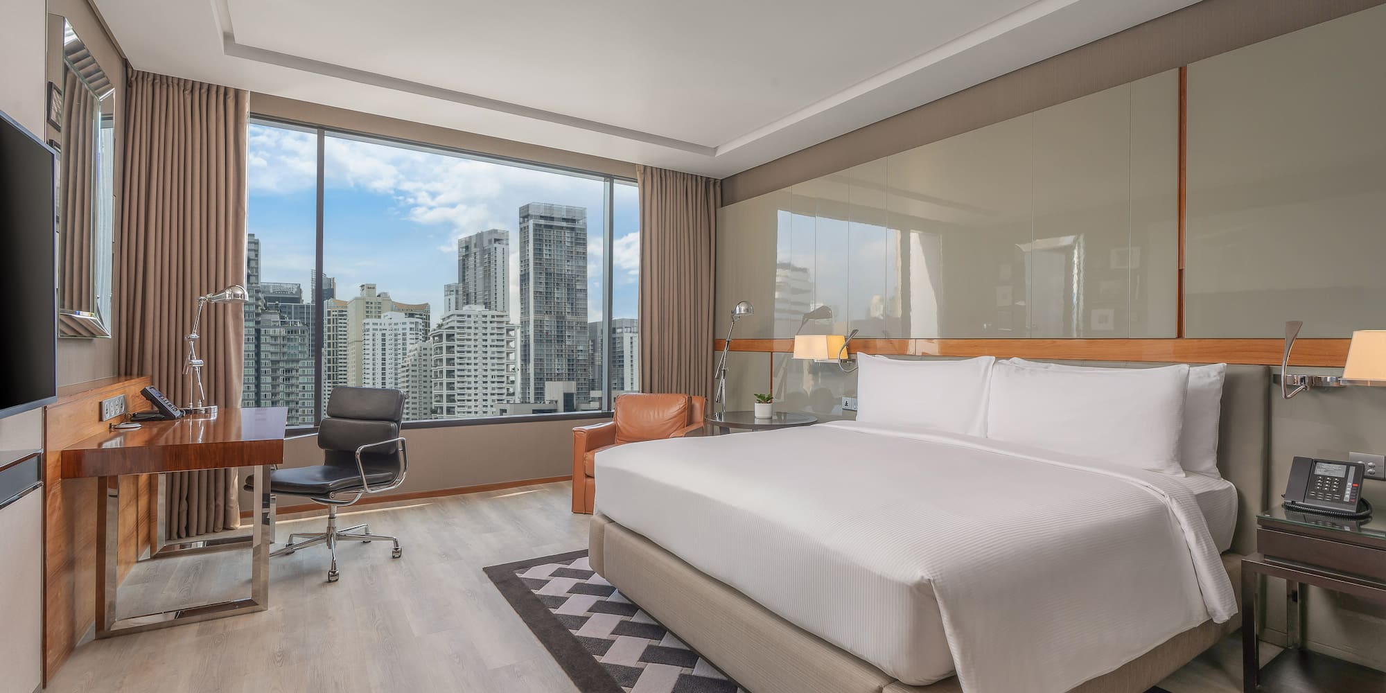 Modern hotel bedroom with large bed, work desk, and a city skyline view.