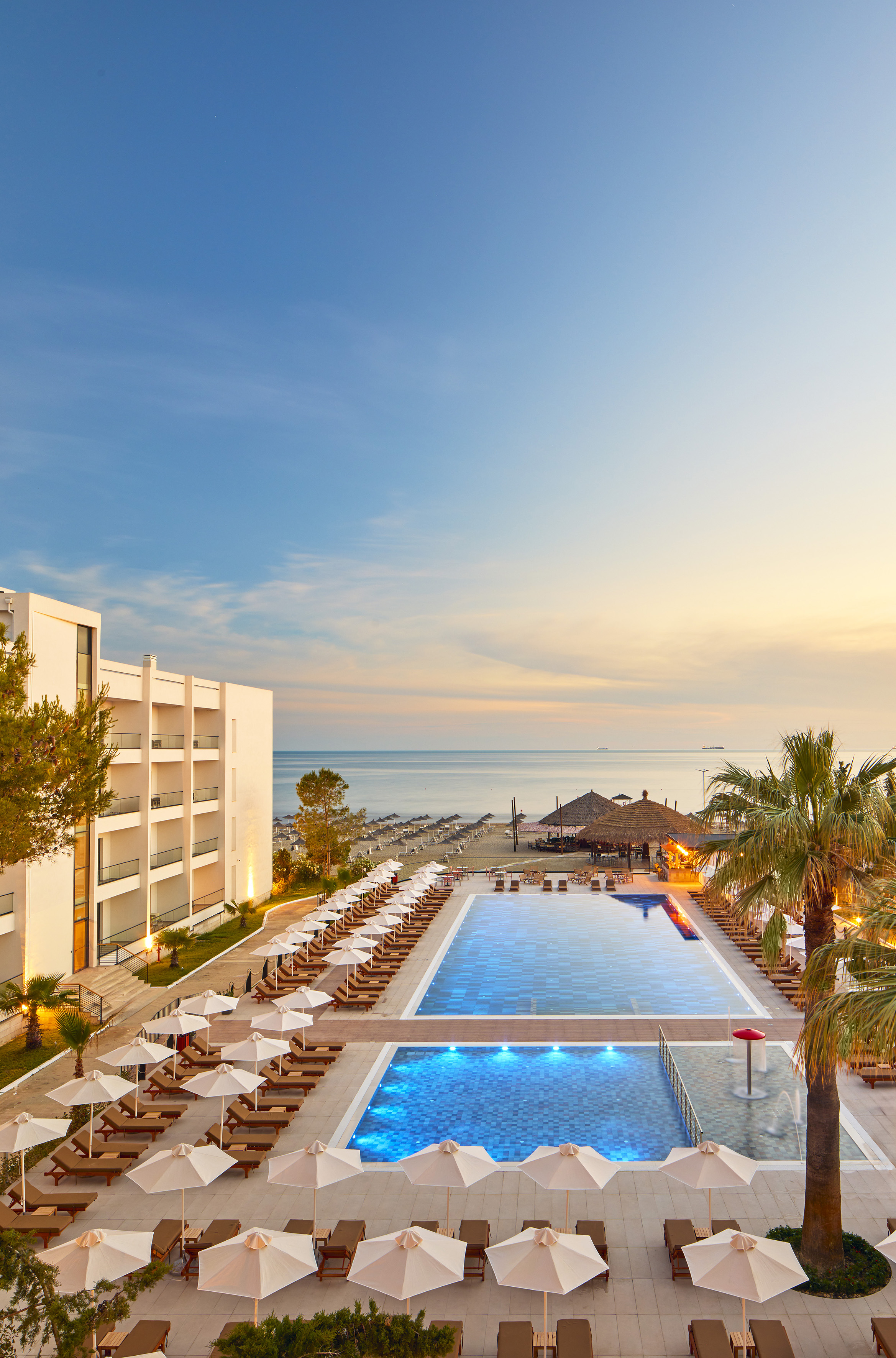 Beach resort with illuminated pools, lounge chairs, and ocean at sunset.