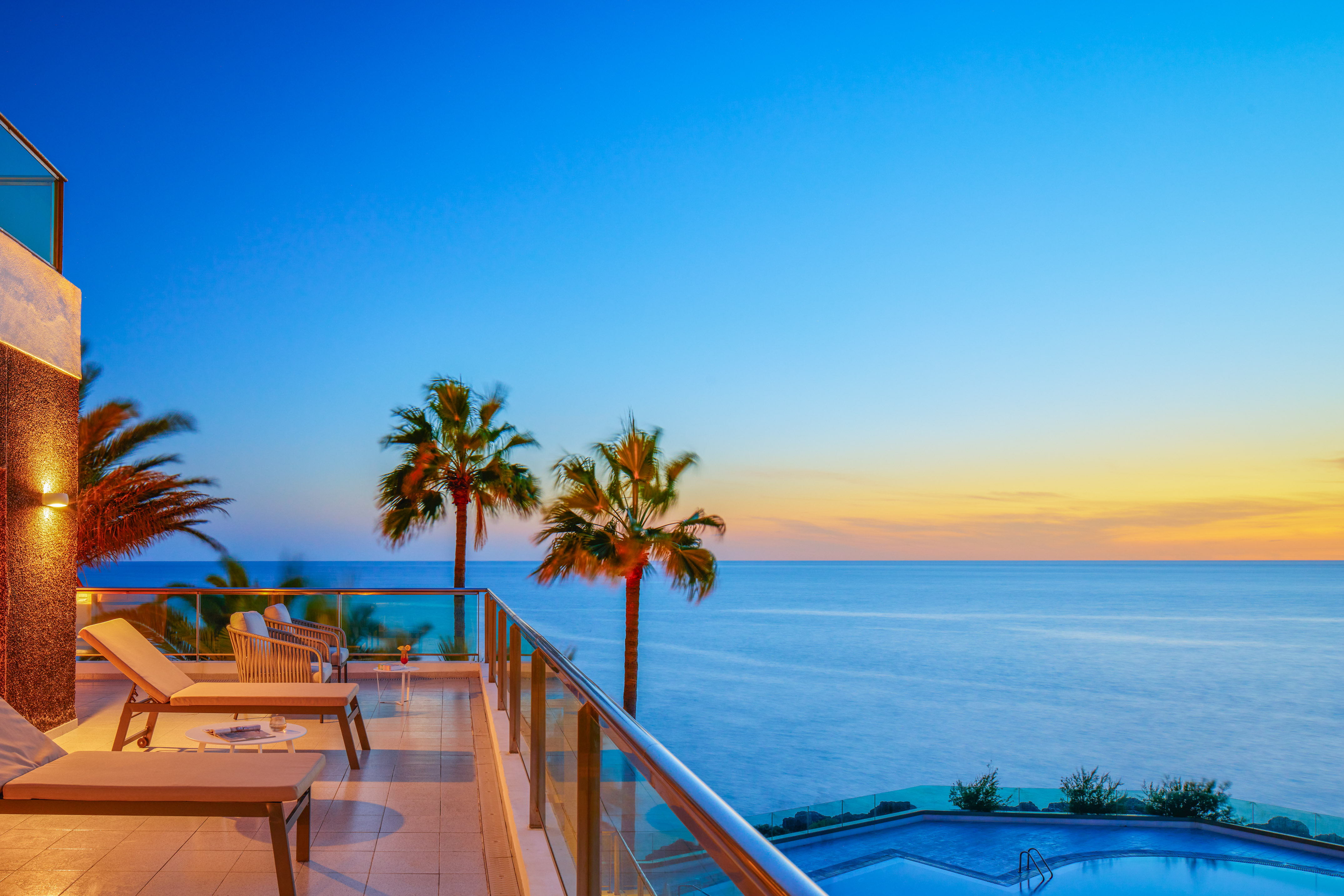 Luxury ocean view balcony with palm trees, sunset sky, and resort pool.