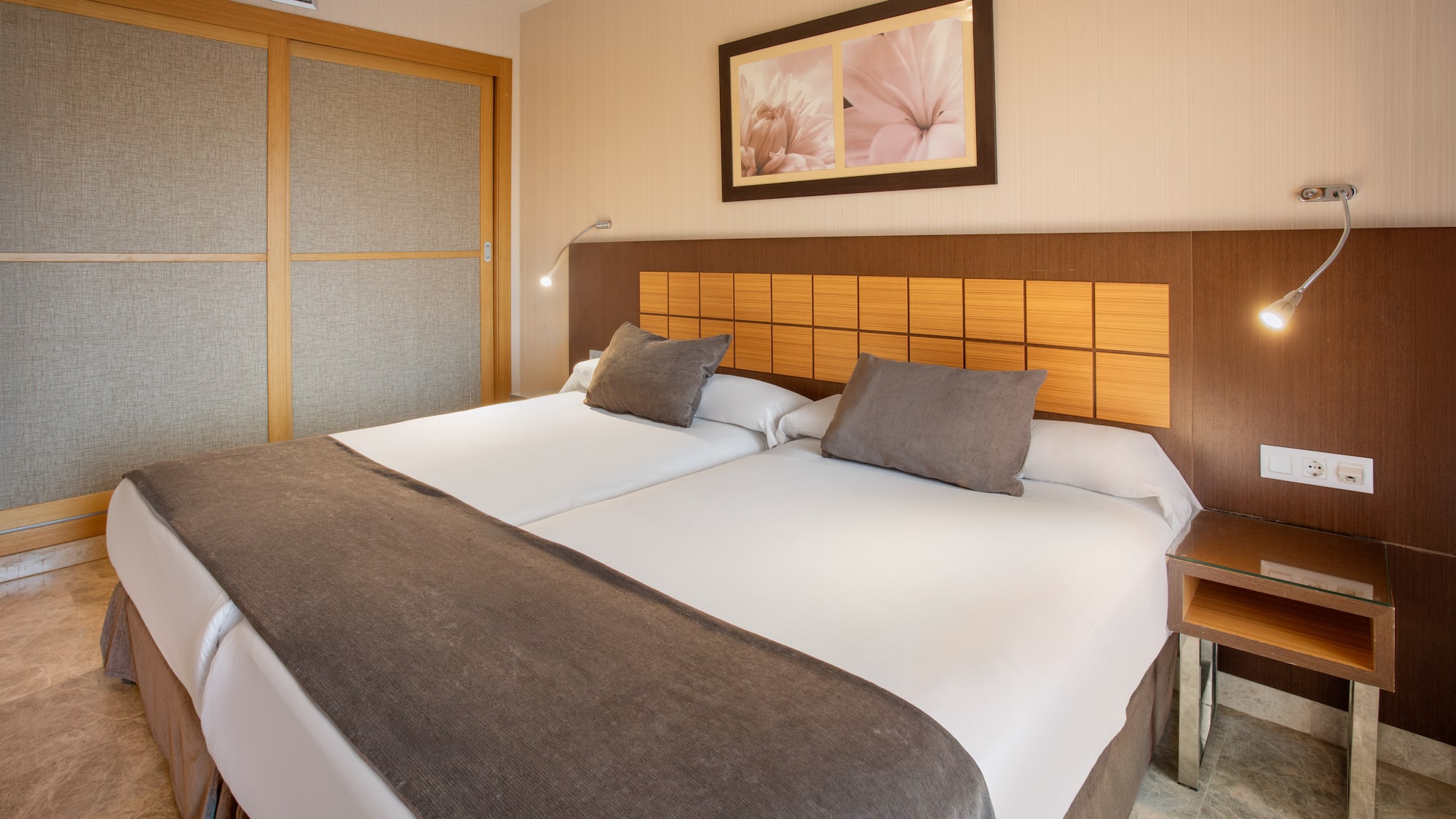 Modern hotel room with twin beds, brown headboard, and sliding wardrobe.