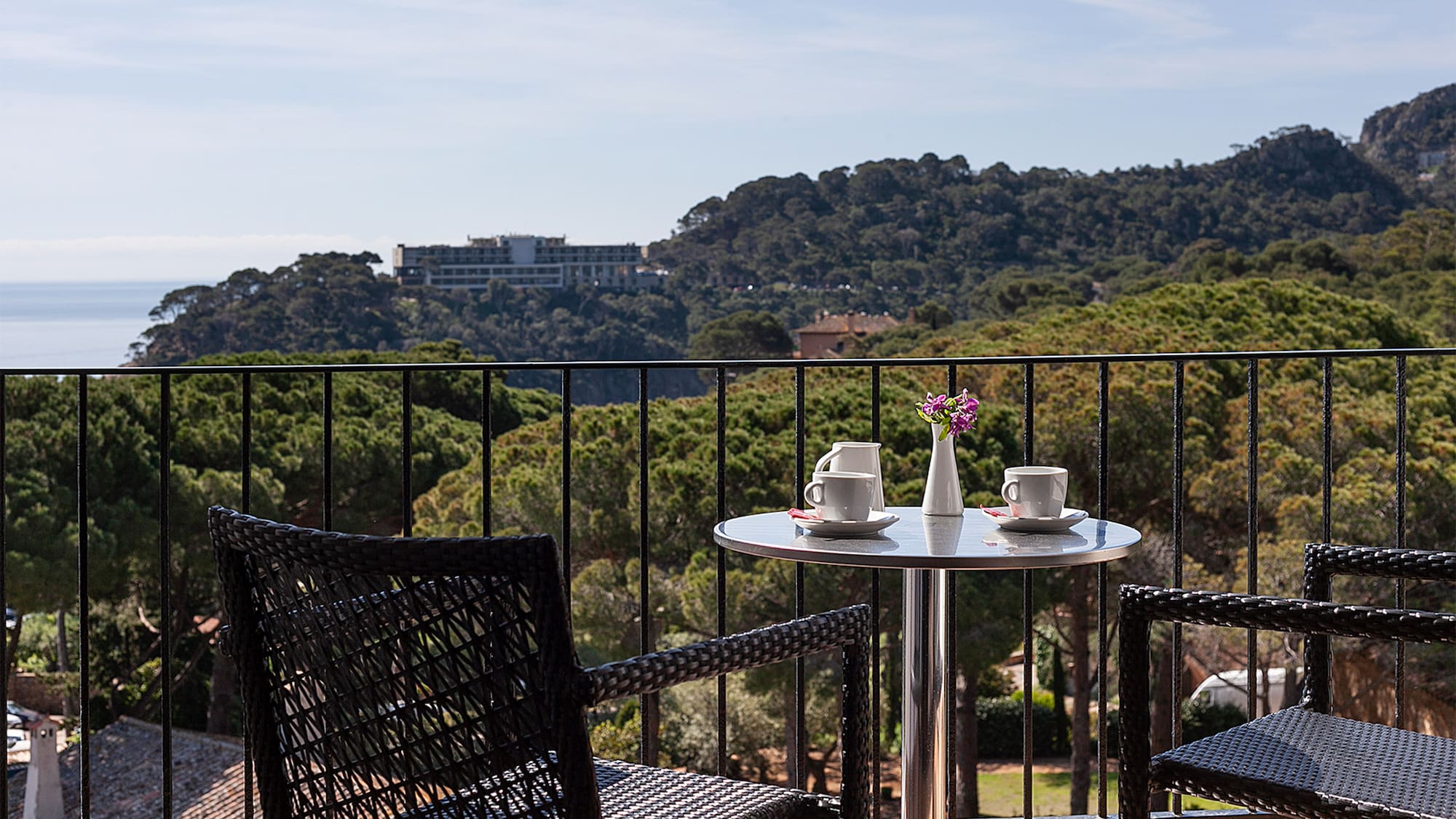 Balcony with coffee on table, chairs, overlooking sea and forest.