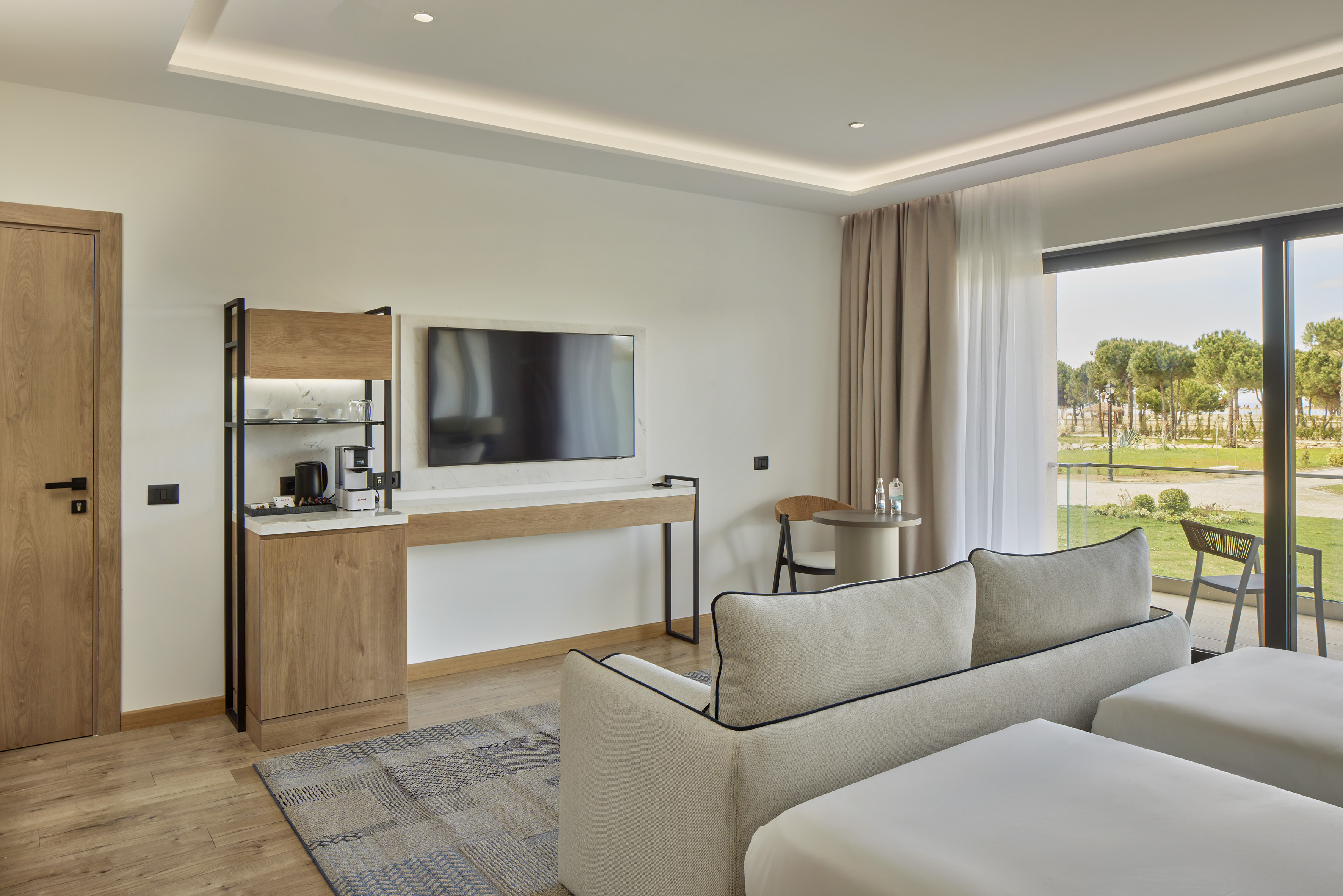 Modern hotel room with TV, sofa, minibar, and balcony garden view.