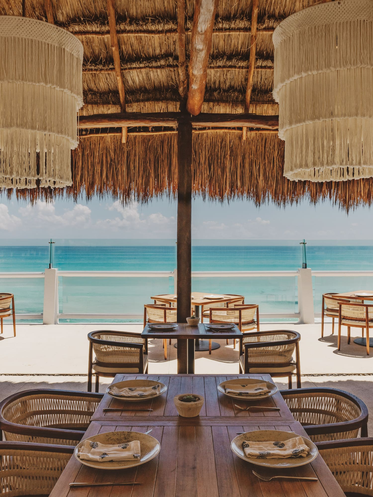 Oceanfront restaurant with thatched roof, macrame chandeliers, and outdoor tables.