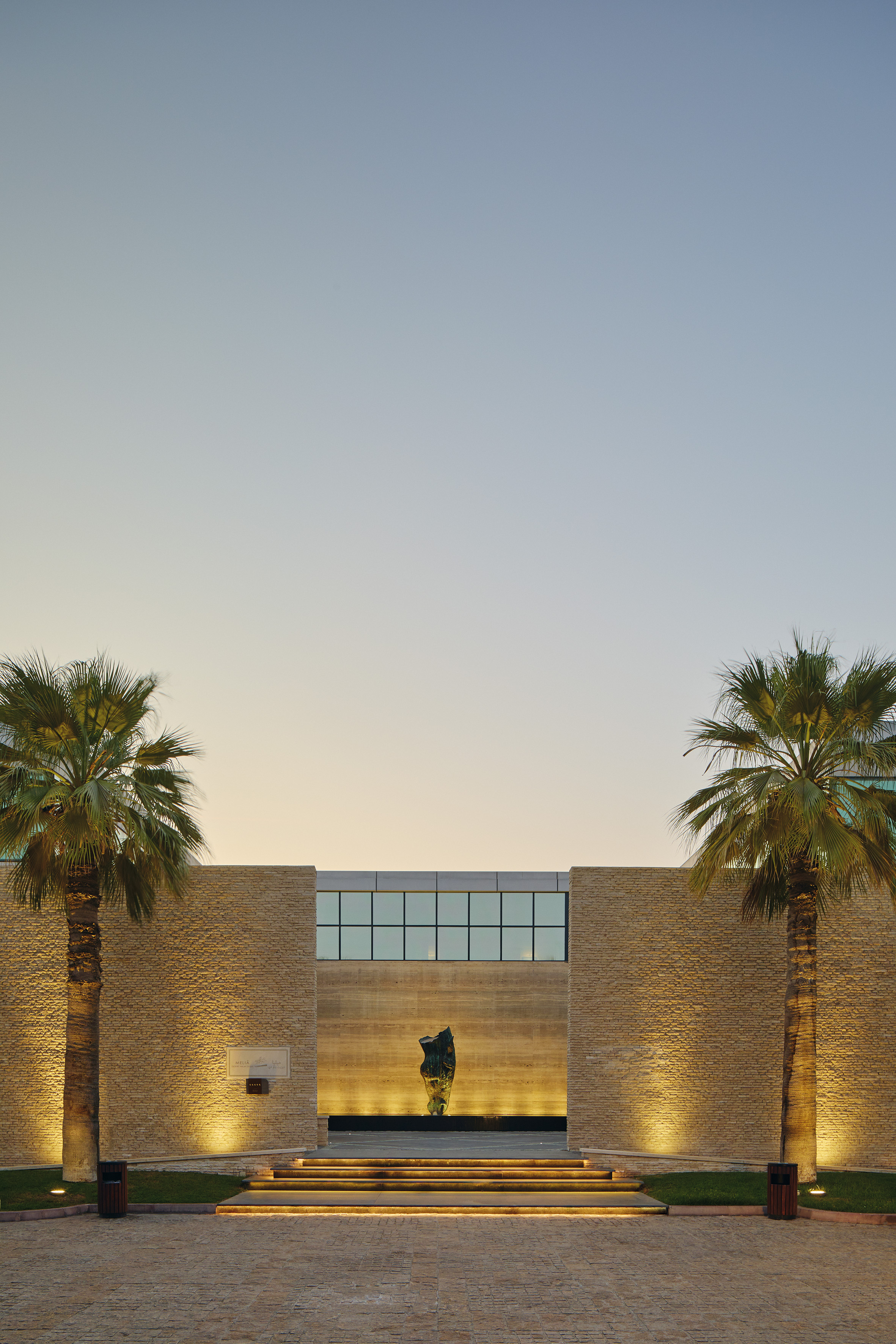 Modern hotel entrance, illuminated steps, palm trees, and abstract sculpture.