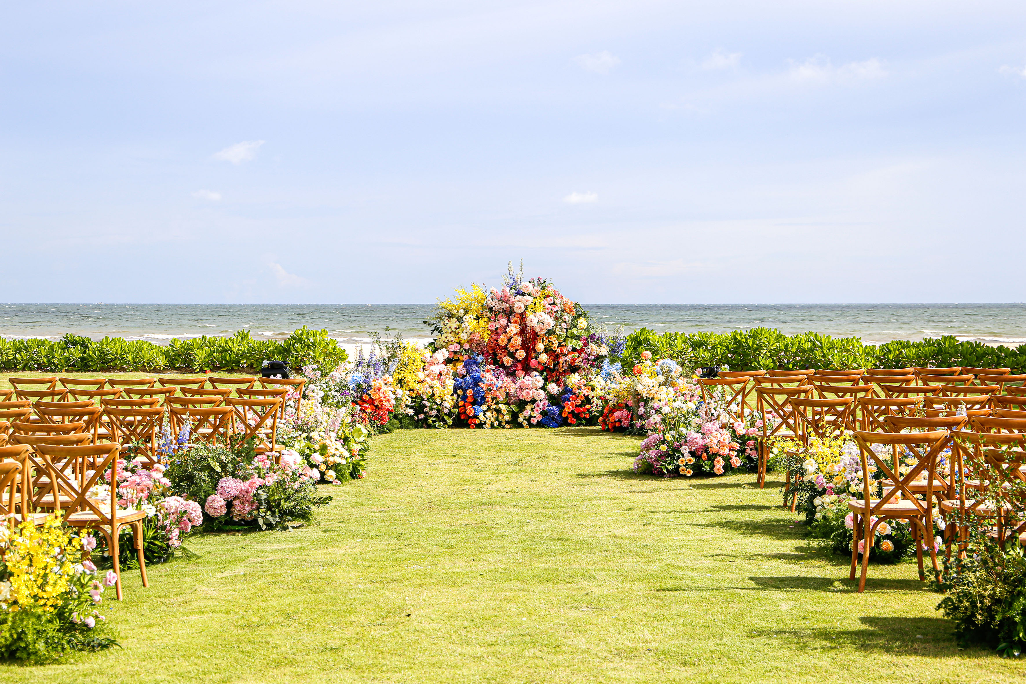 Vibrant floral beach wedding arch, chairs on grass, ocean background.