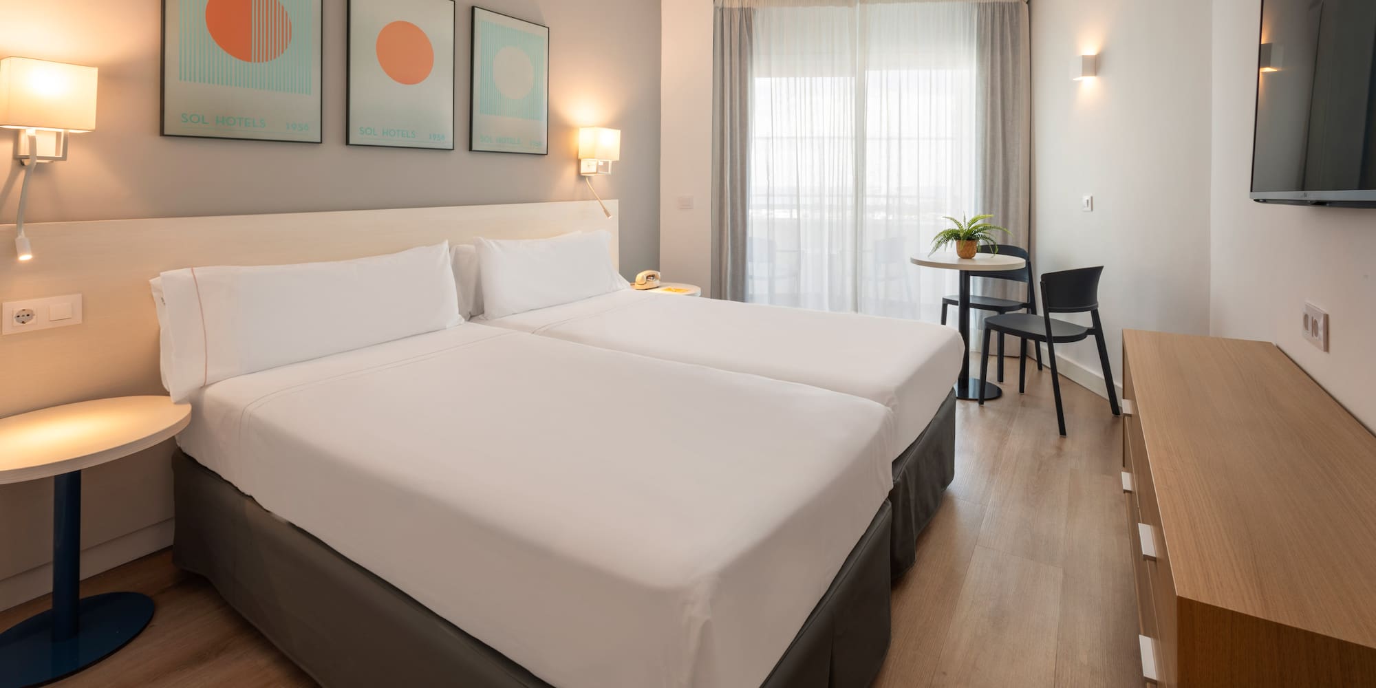 Bright modern hotel room with twin beds, seating area, and wall-mounted TV.