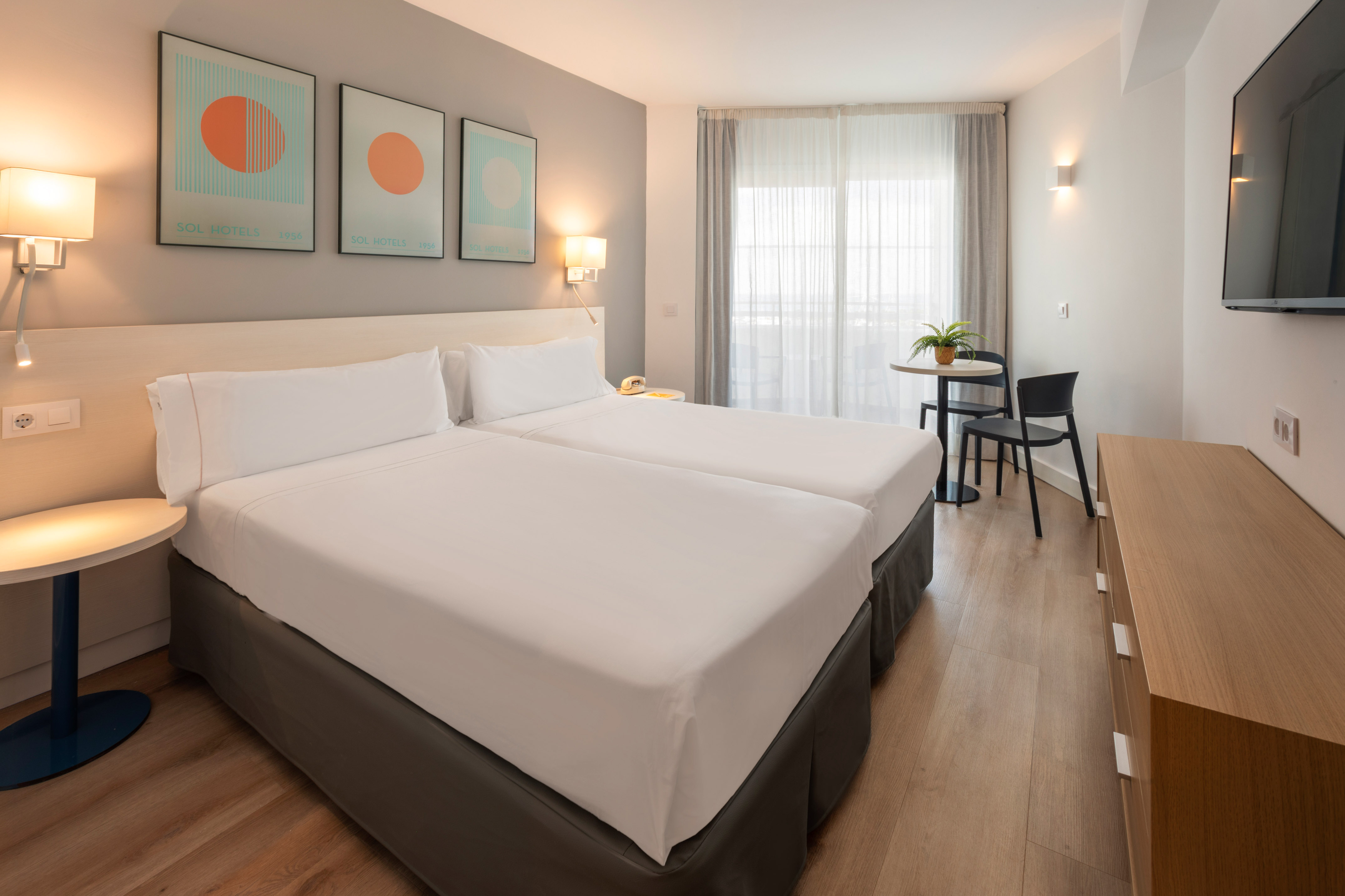 Bright modern hotel room with twin beds, seating area, and wall-mounted TV.