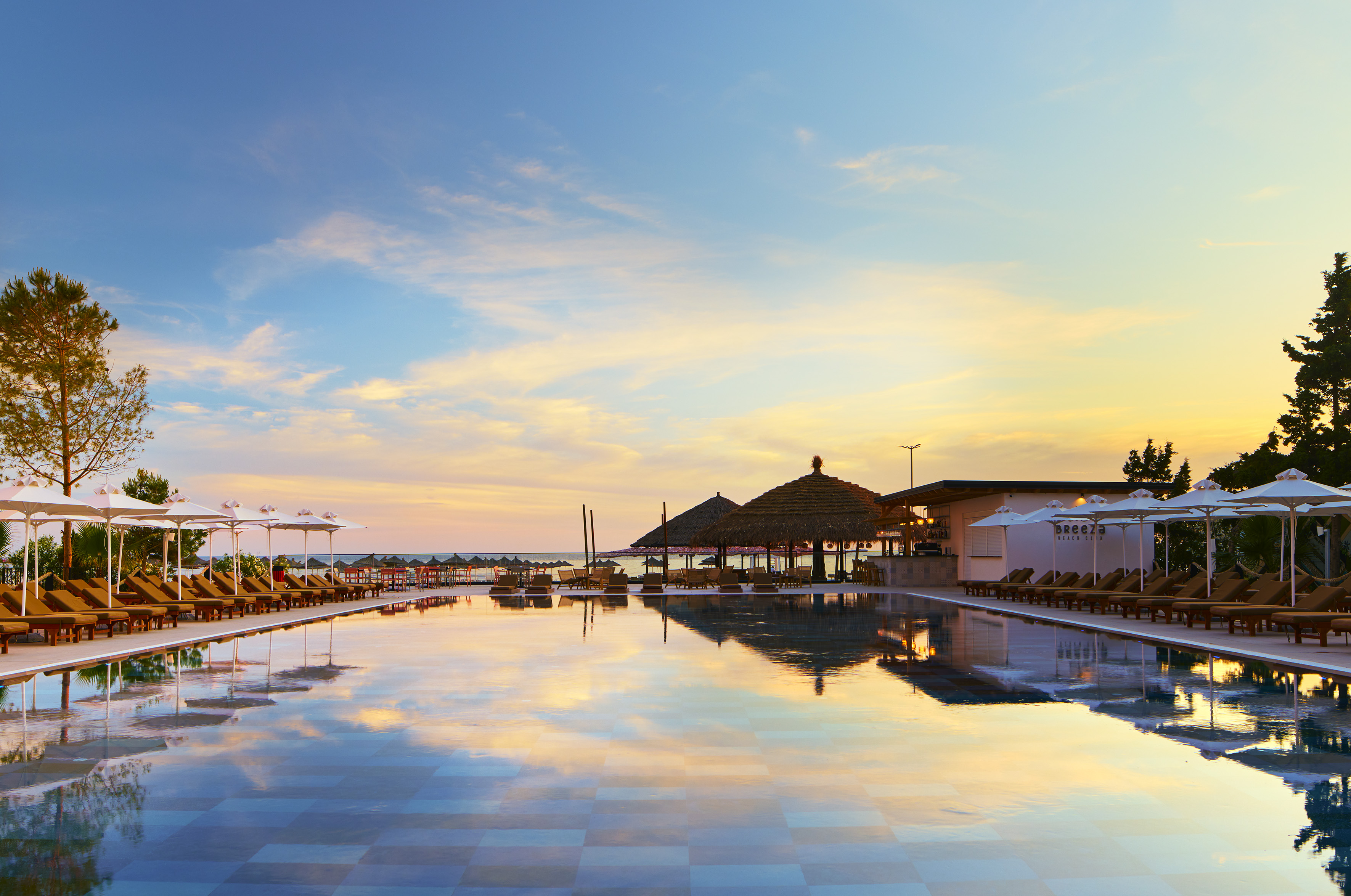 Resort swimming pool reflecting sunset sky, surrounded by lounge chairs and beach club.