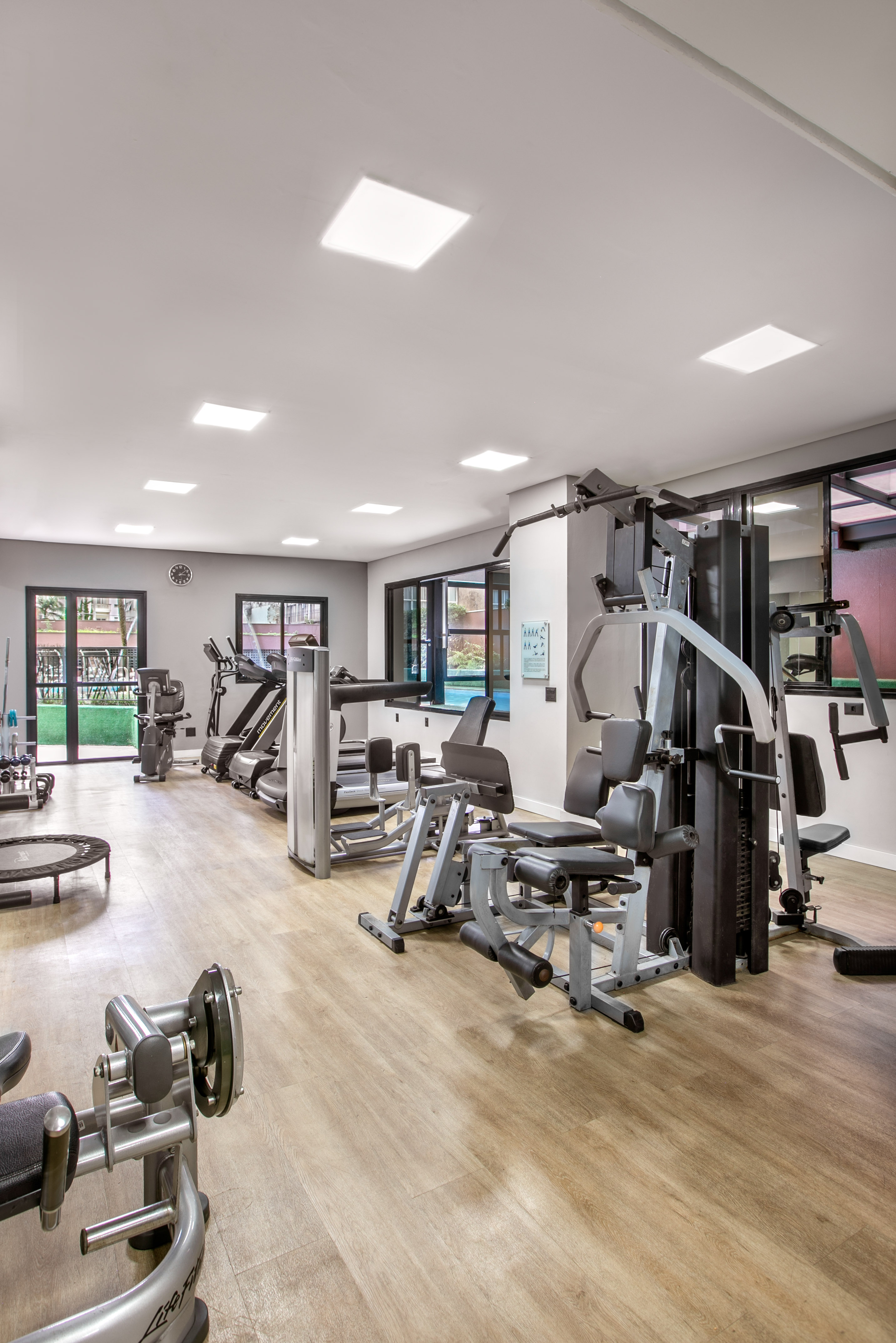 Modern gym with various exercise equipment, light wood floor, and large windows.