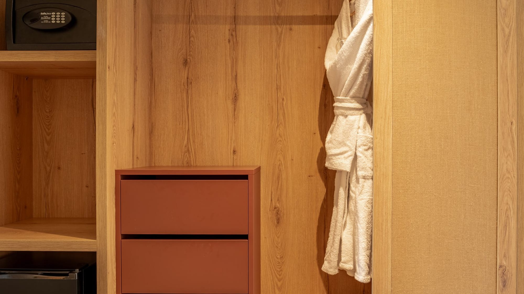 Wooden hotel wardrobe with built-in safe, minibar, drawers, and white bathrobe.
