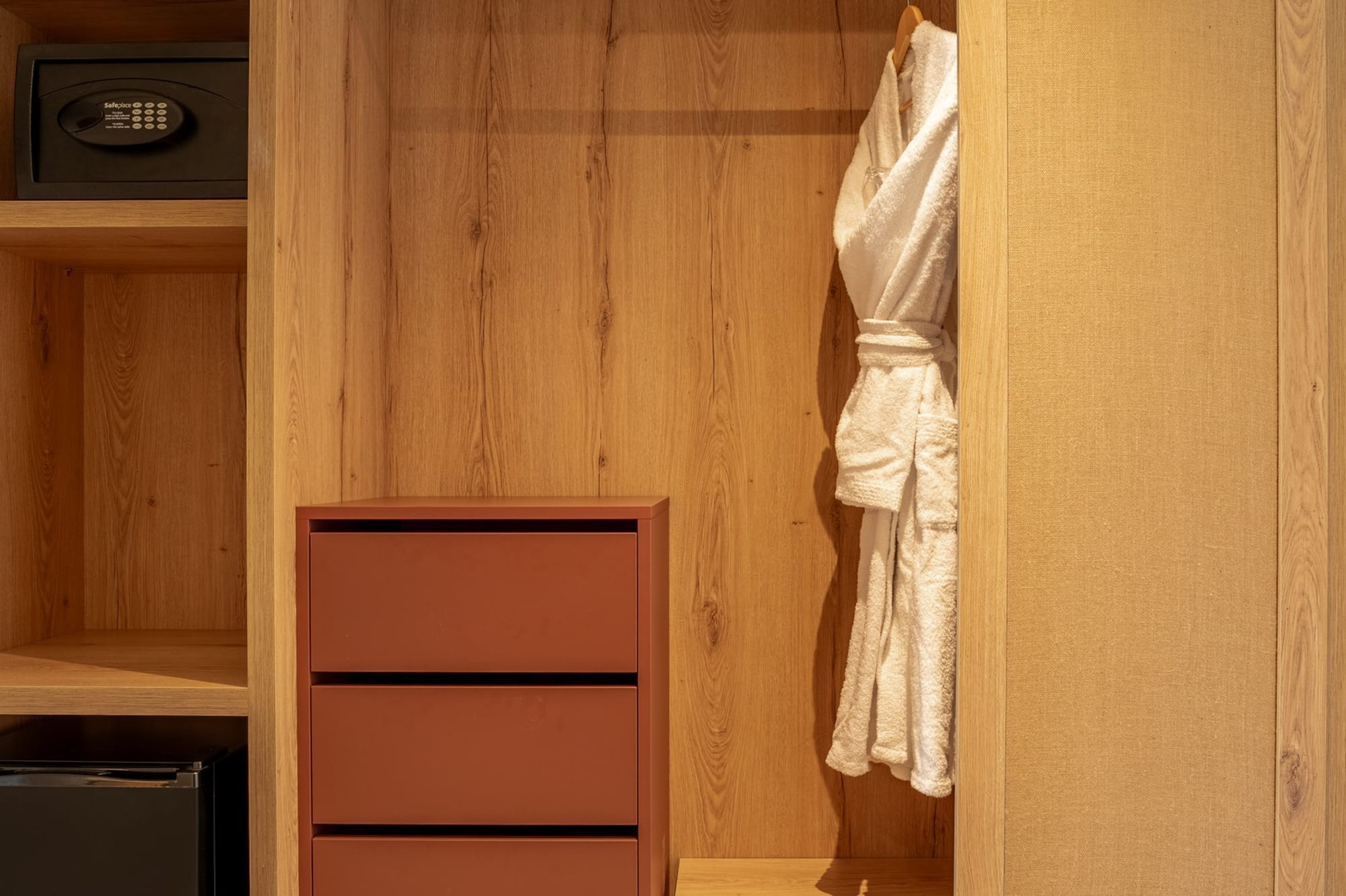 Wooden hotel wardrobe with built-in safe, minibar, drawers, and white bathrobe.