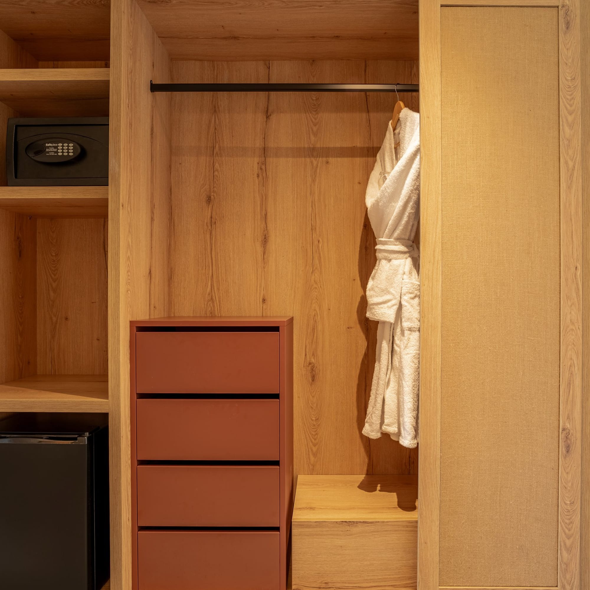 Wooden hotel wardrobe with built-in safe, minibar, drawers, and white bathrobe.