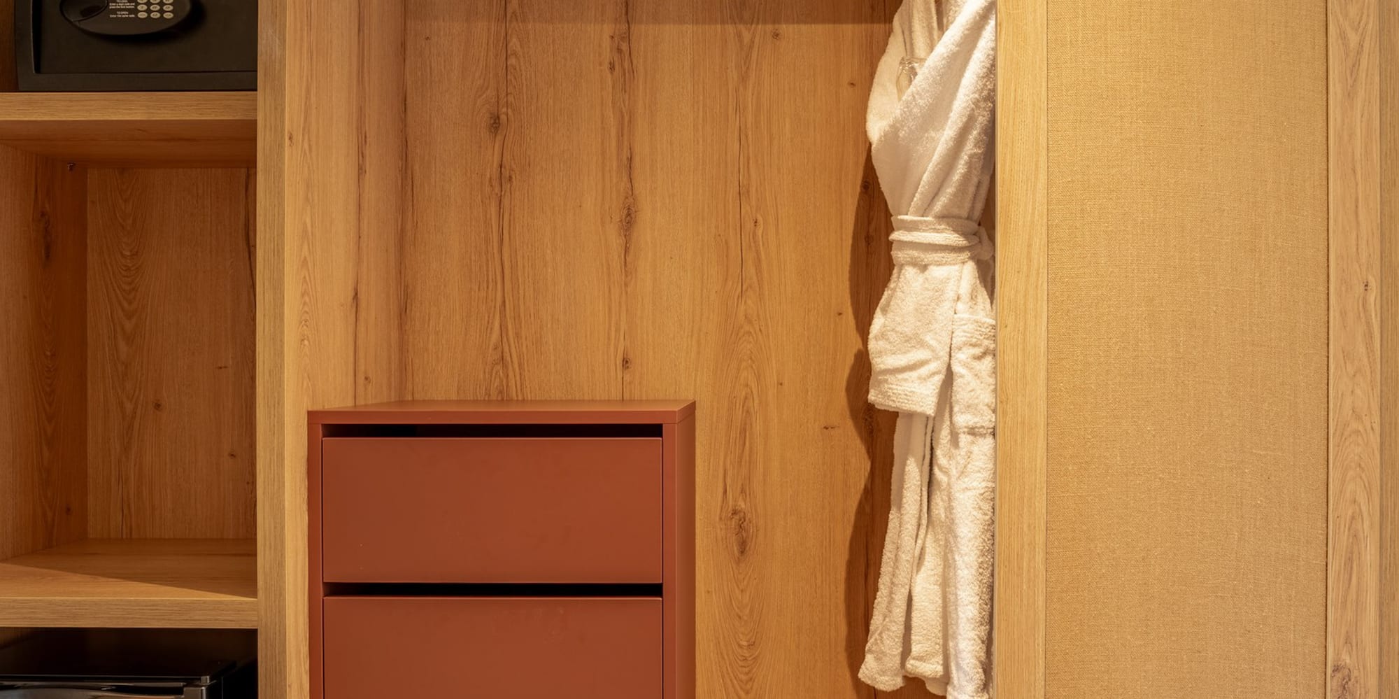 Wooden hotel wardrobe with built-in safe, minibar, drawers, and white bathrobe.