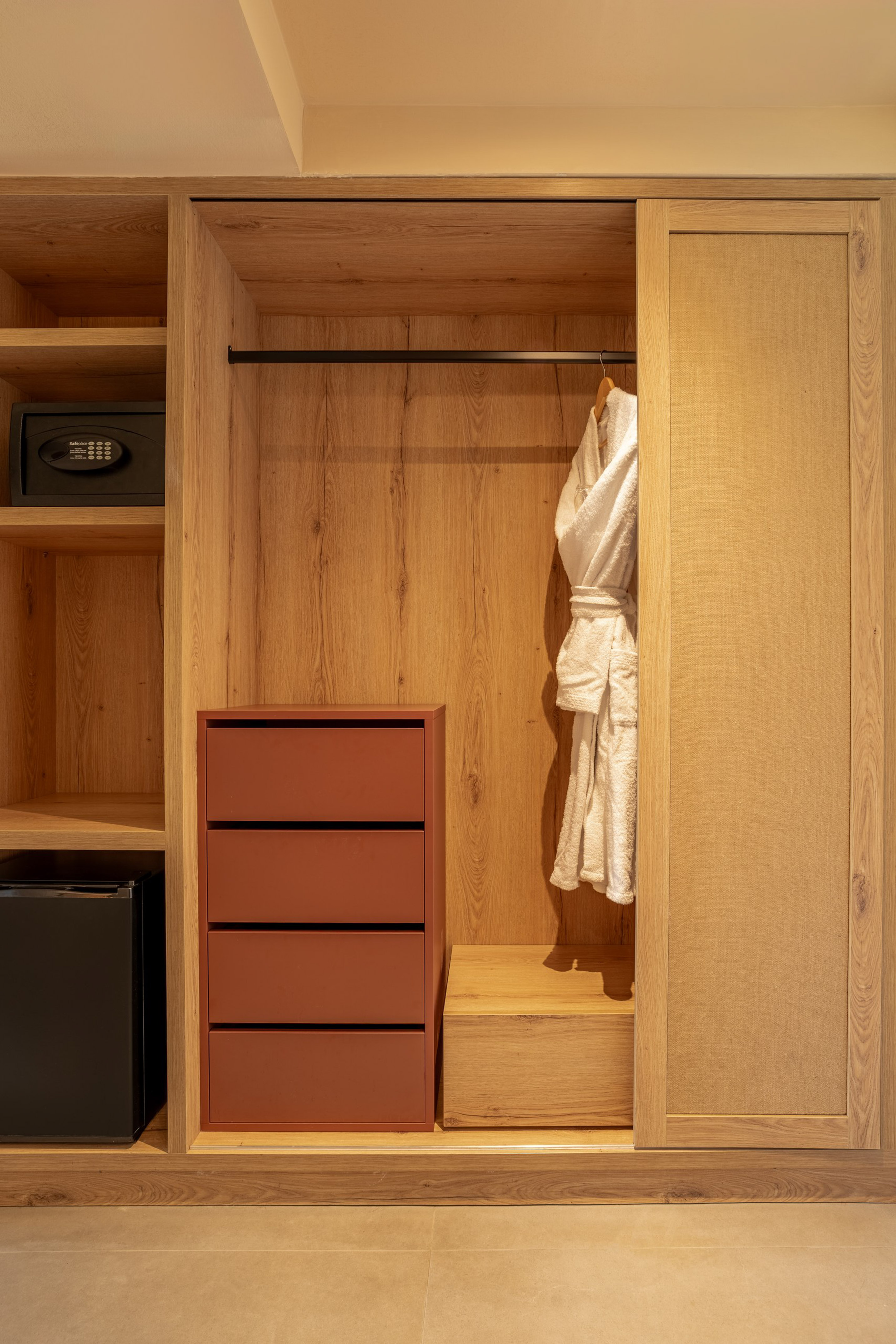 Wooden hotel wardrobe with built-in safe, minibar, drawers, and white bathrobe.