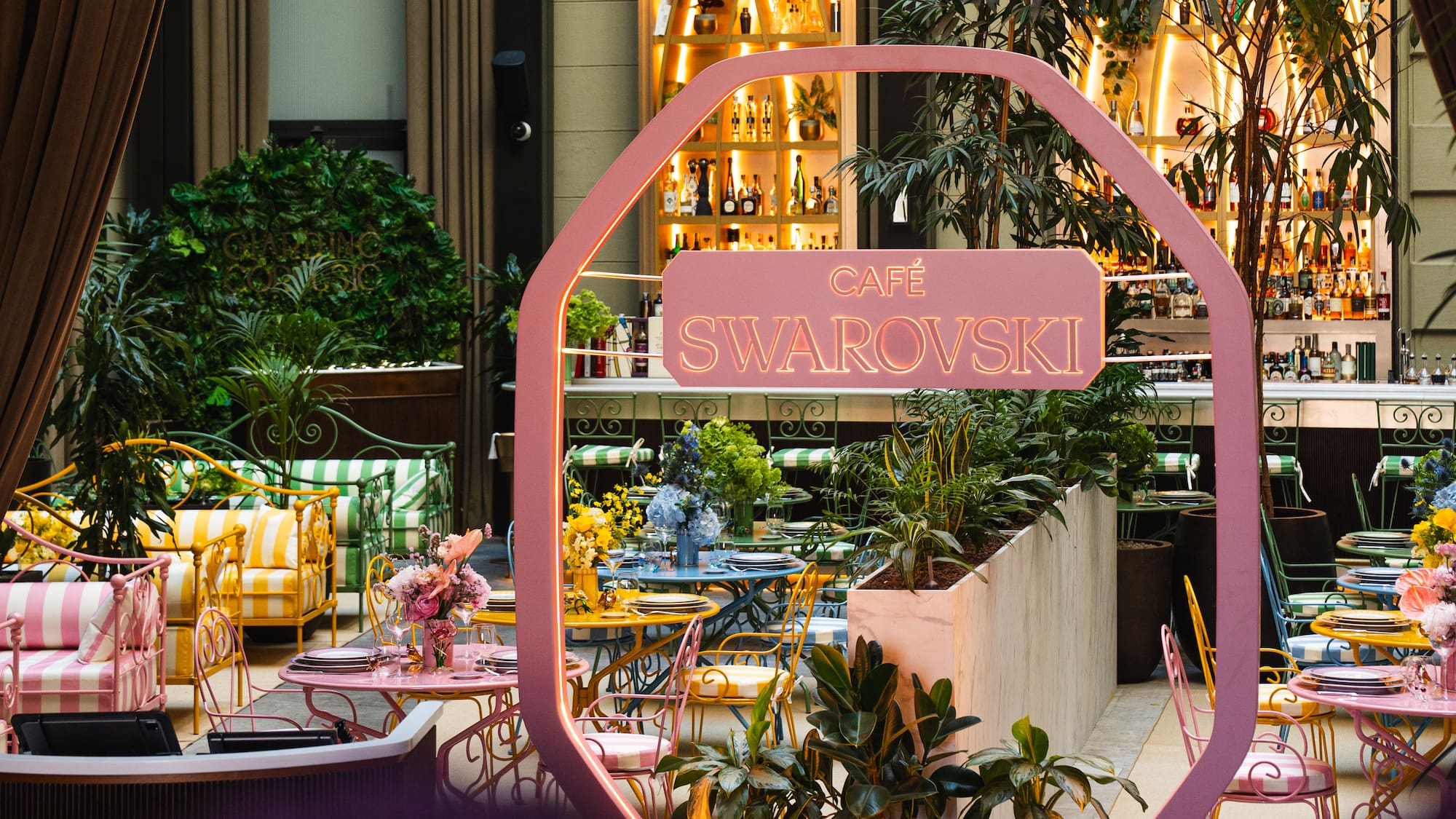 Vibrant Cafe Swarovski with colorful seating, lush plants, and a glowing bar.