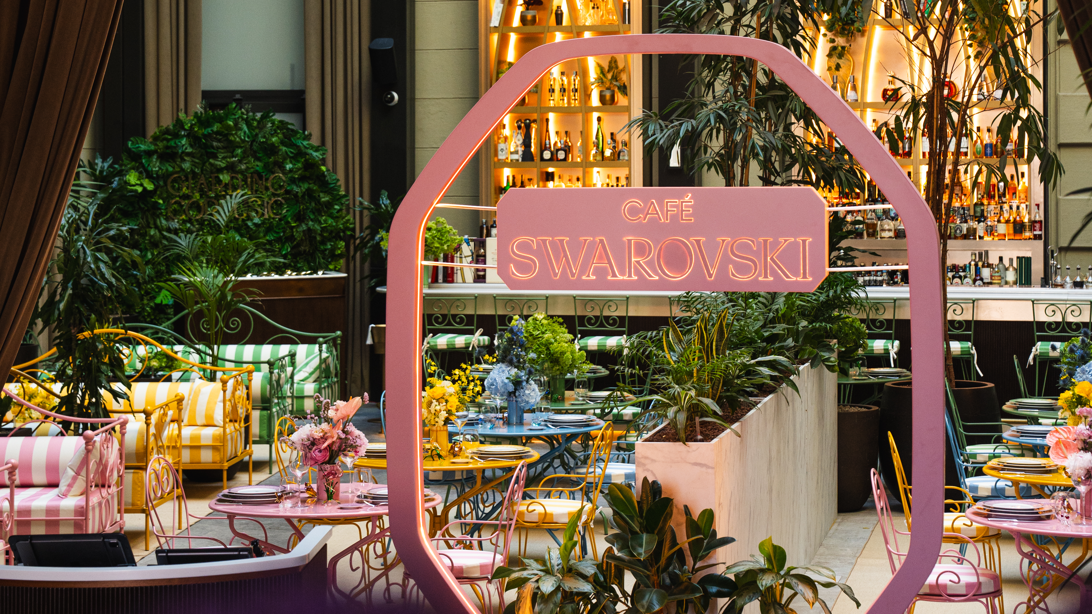 Vibrant Cafe Swarovski with colorful seating, lush plants, and a glowing bar.