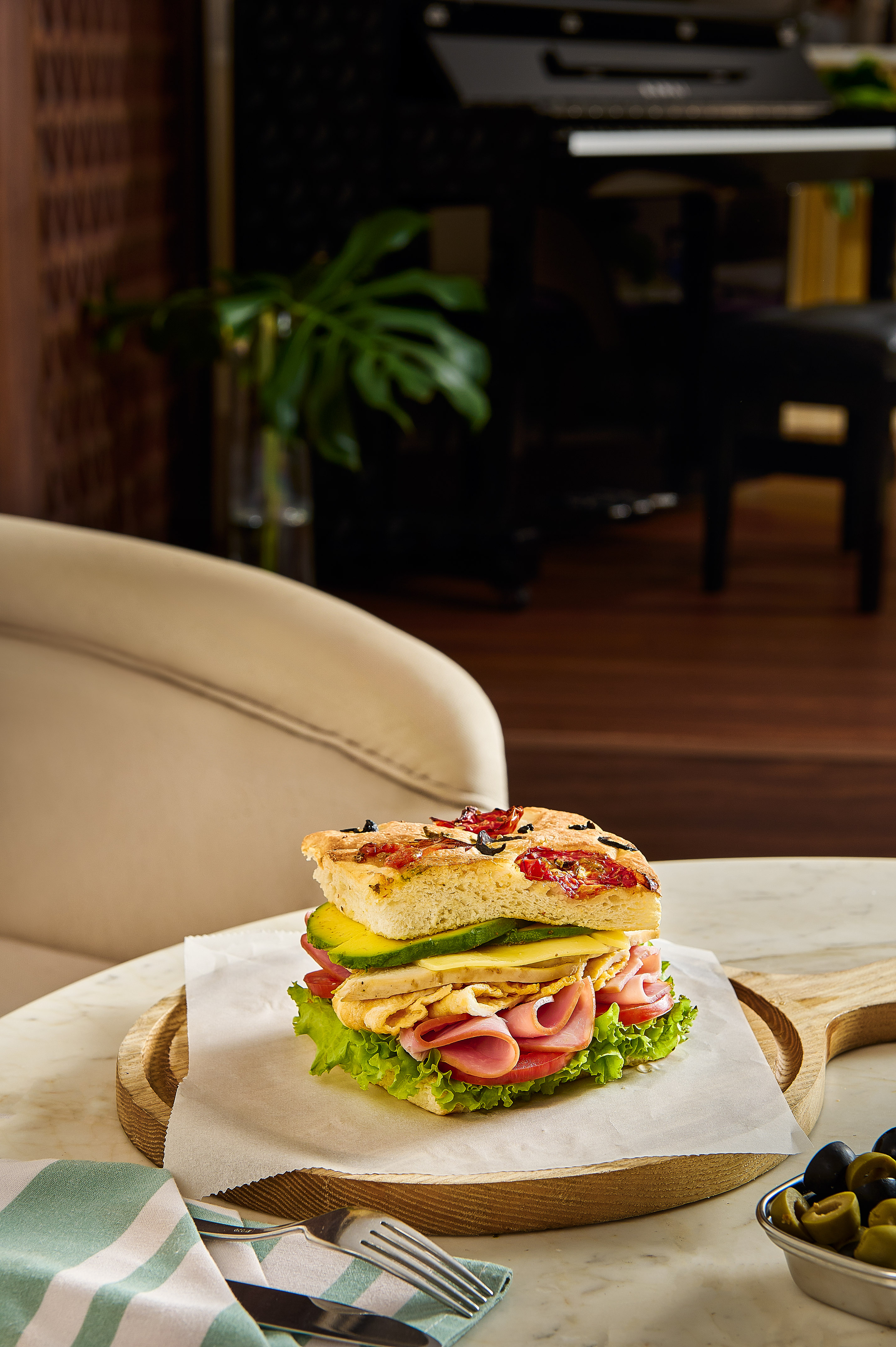 Focaccia sandwich with ham, cheese, avocado, lettuce, and olives on a wooden board.