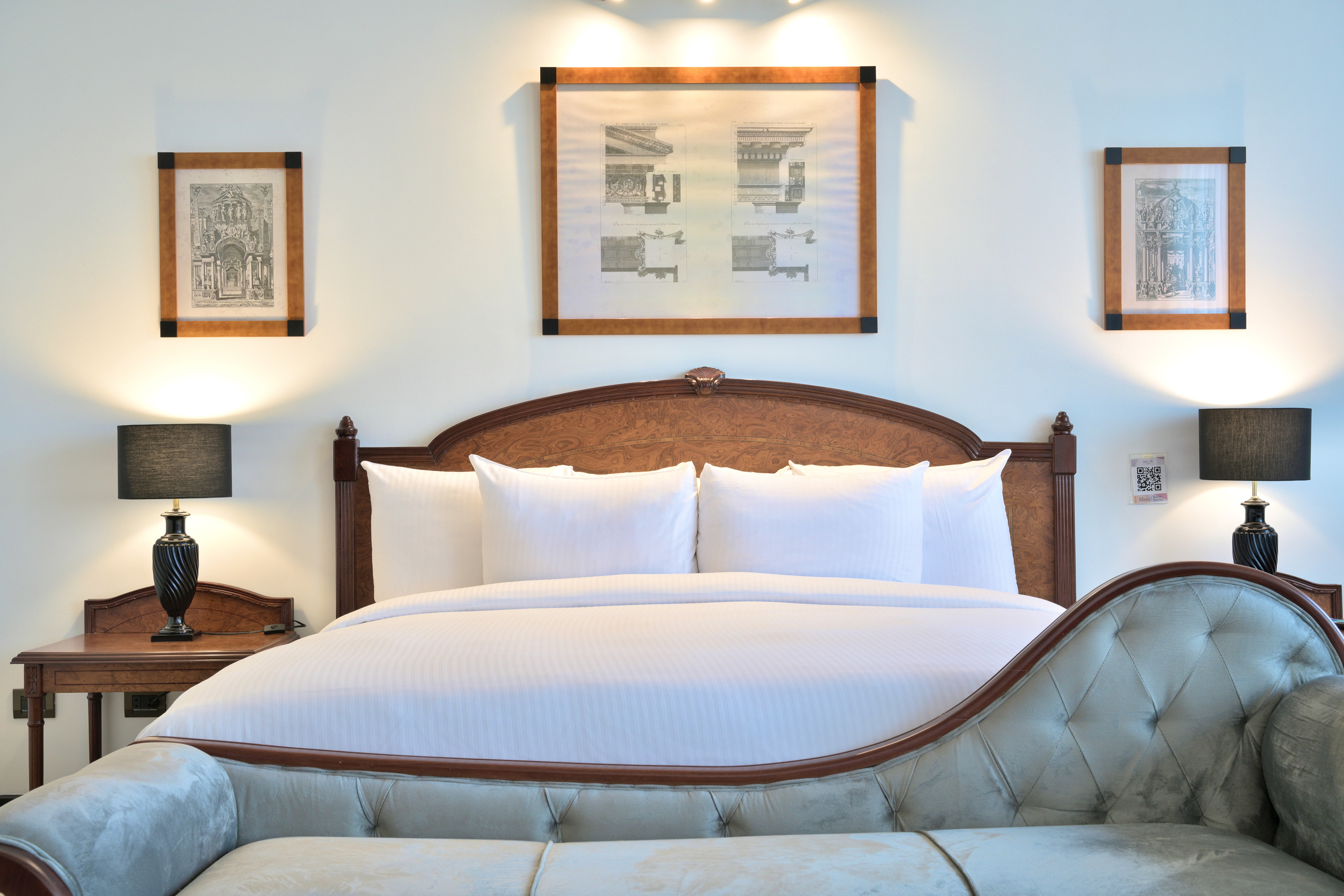 Luxurious hotel bedroom with white bed, wooden headboard, lamps, and chaise lounge.