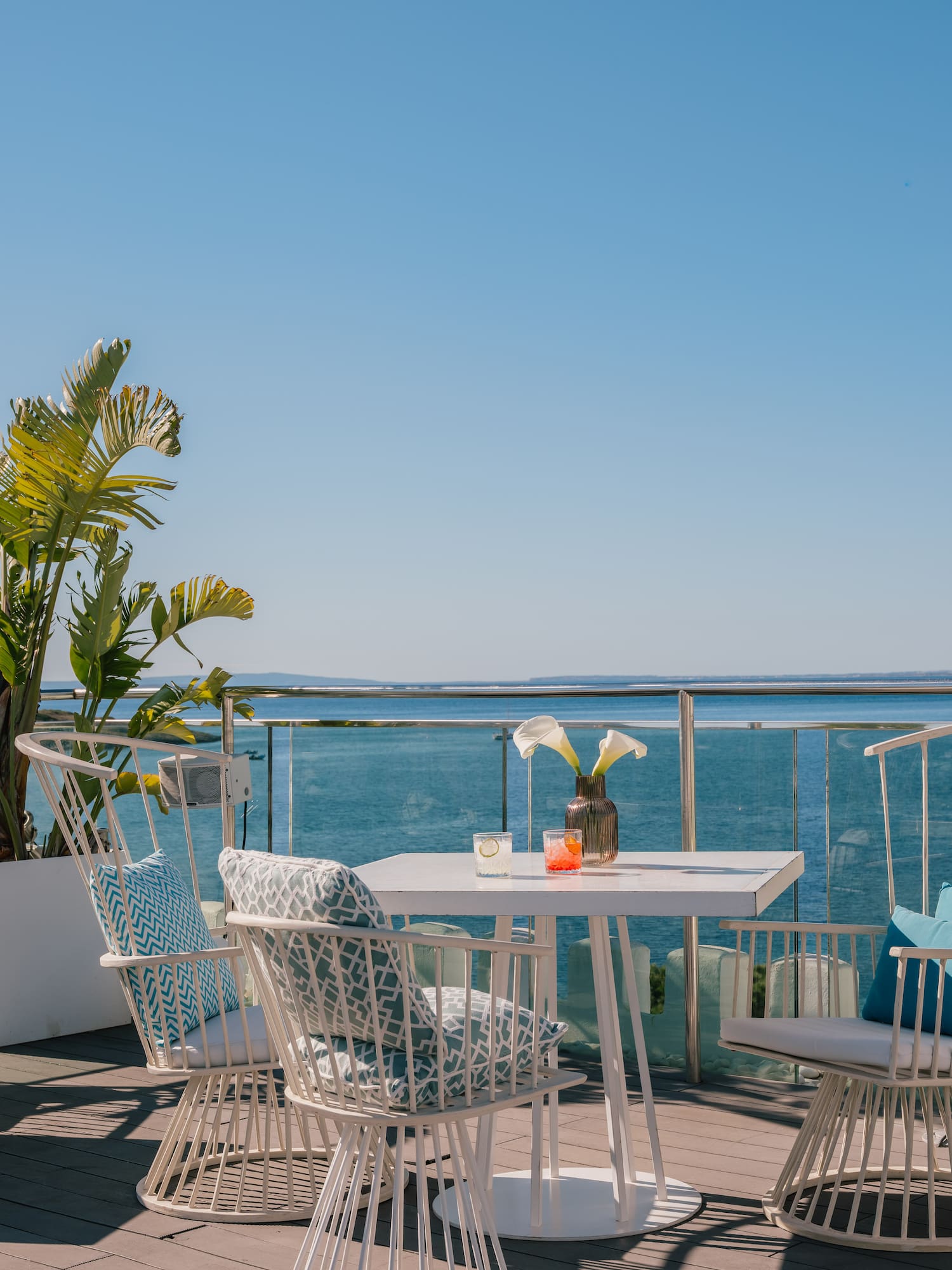 Outdoor terrace with white dining set, tropical plant, drinks, and ocean view.