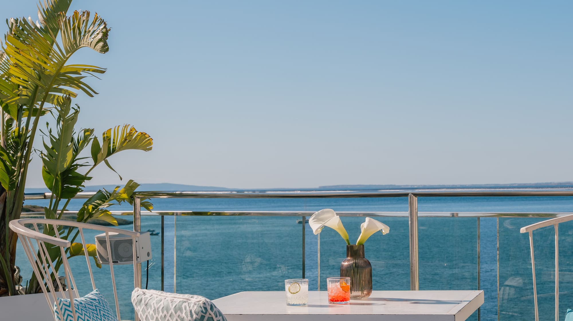 Oceanfront balcony with white patio furniture, drinks, and tropical plant.