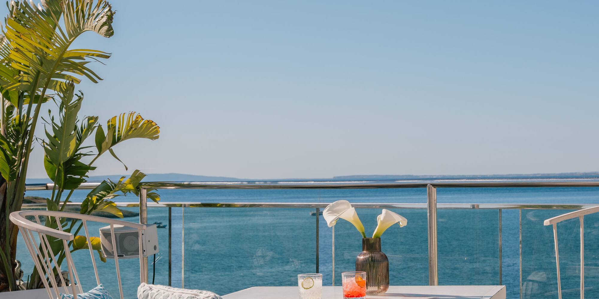 Oceanfront balcony with white patio furniture, drinks, and tropical plant.
