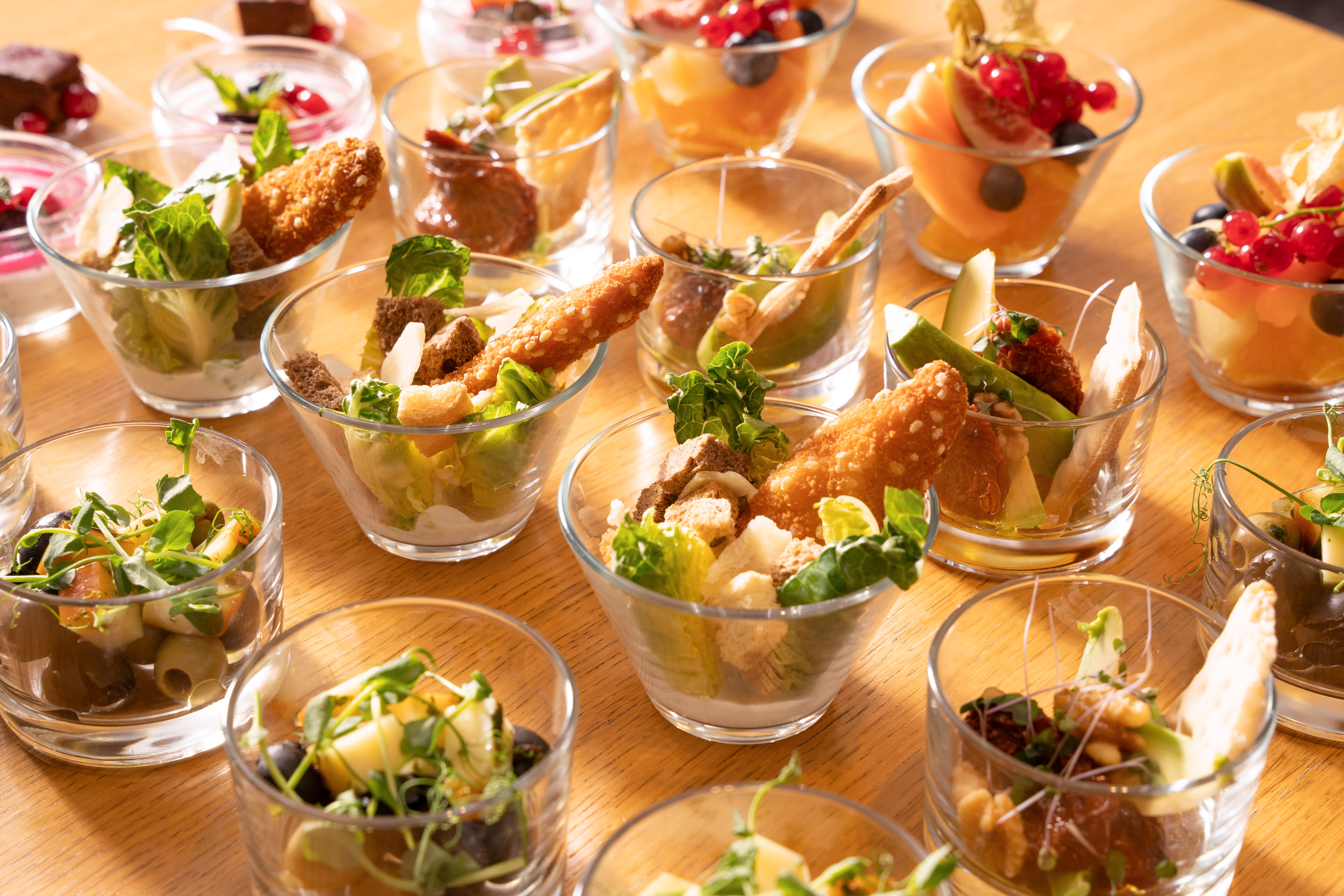 Mini buffet appetizers, salads, and fruit desserts in glass cups on wood table.