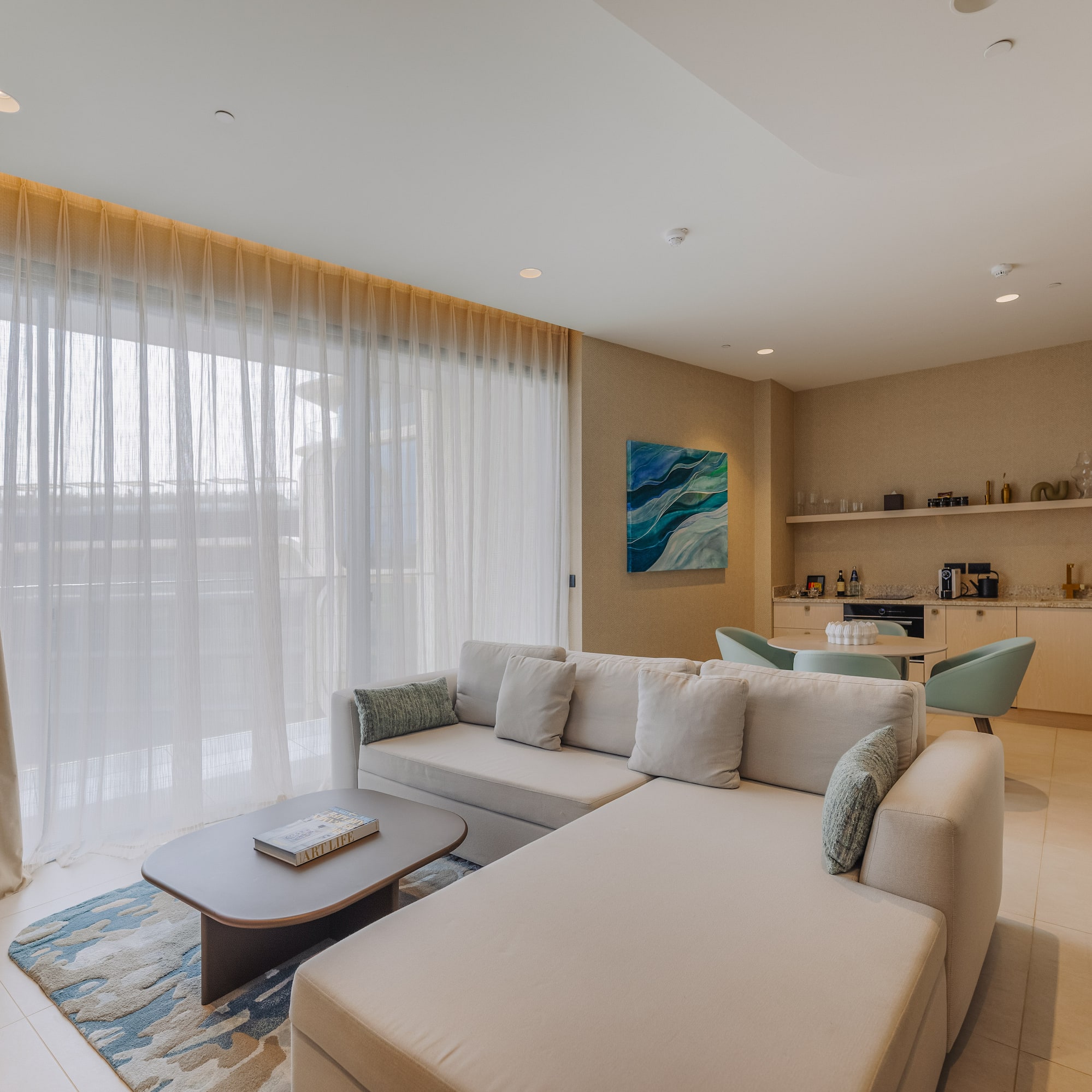 Bright modern living room with large beige sofa, sheer curtains, and small kitchenette.