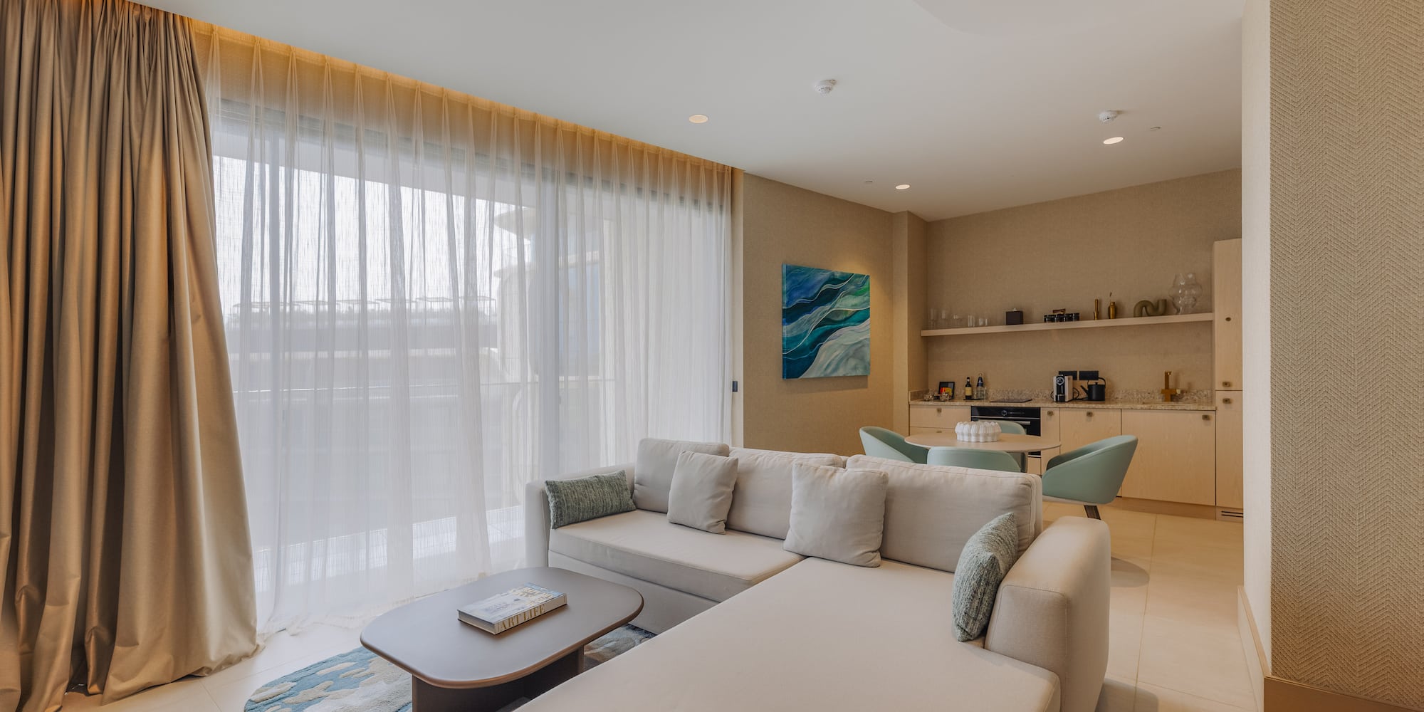 Bright modern living room with large beige sofa, sheer curtains, and small kitchenette.