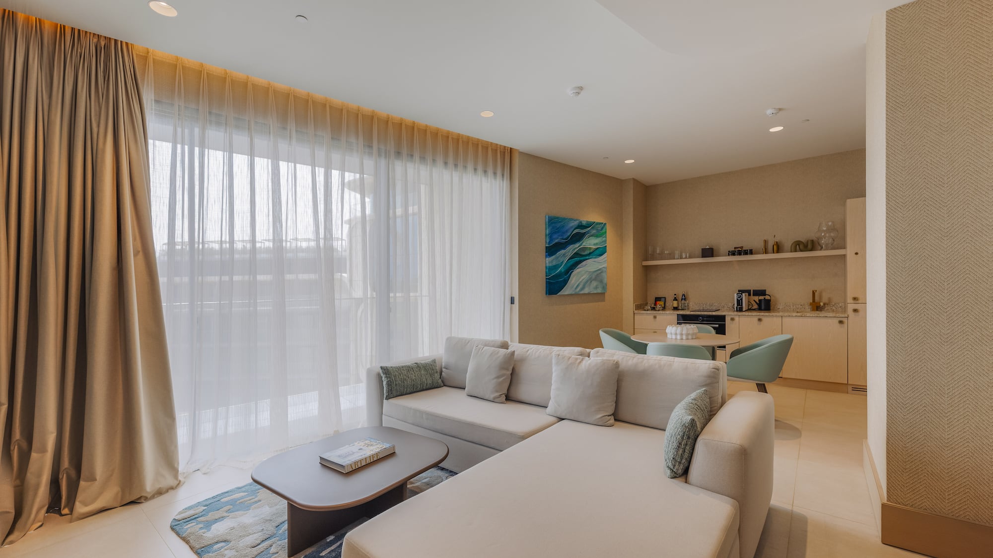 Bright modern living room with large beige sofa, sheer curtains, and small kitchenette.