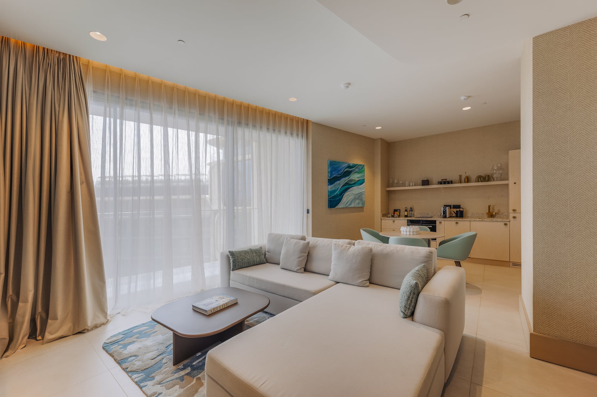 Bright modern living room with large beige sofa, sheer curtains, and small kitchenette.