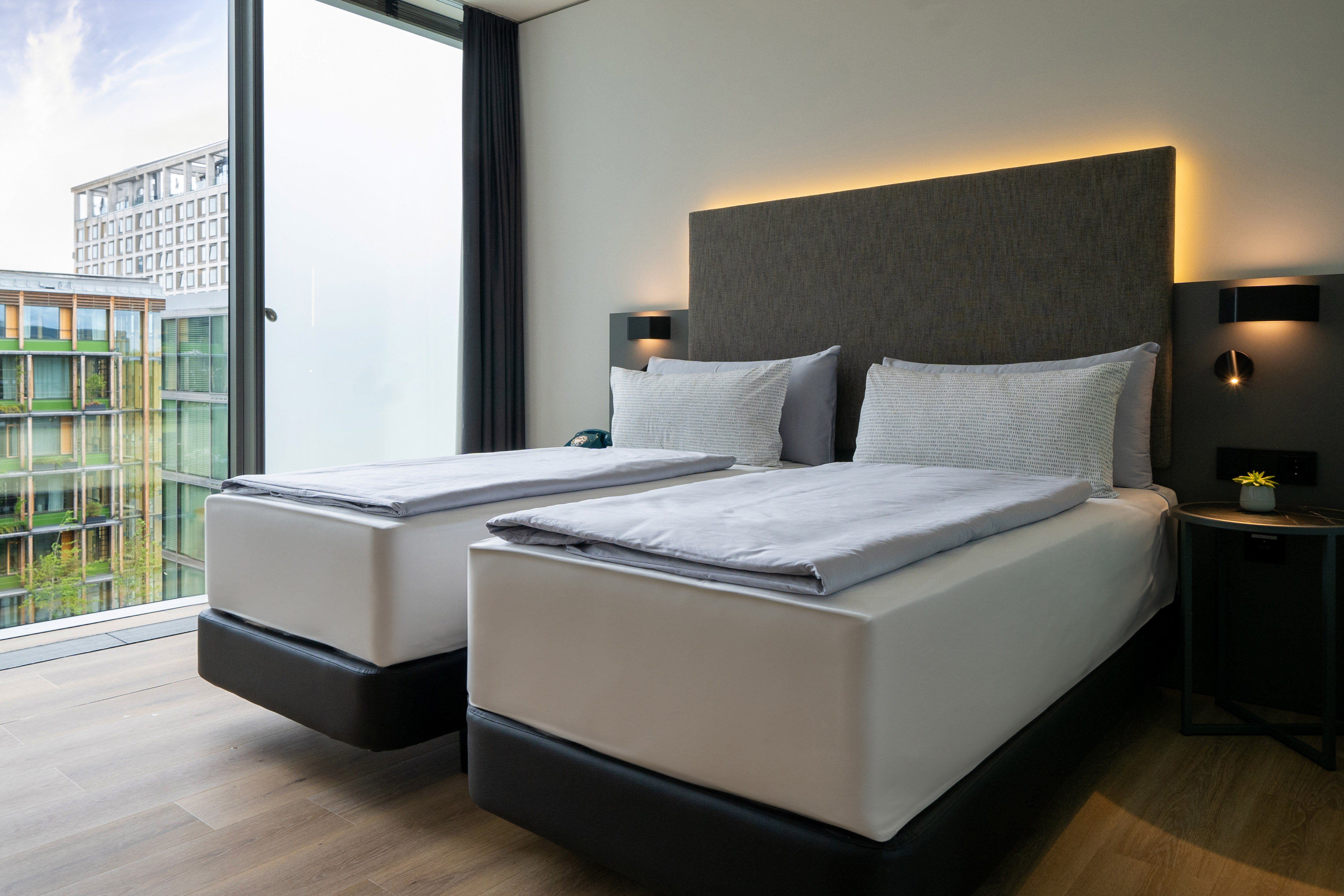 Modern hotel room with two twin beds, backlit headboard, large window, and city view.