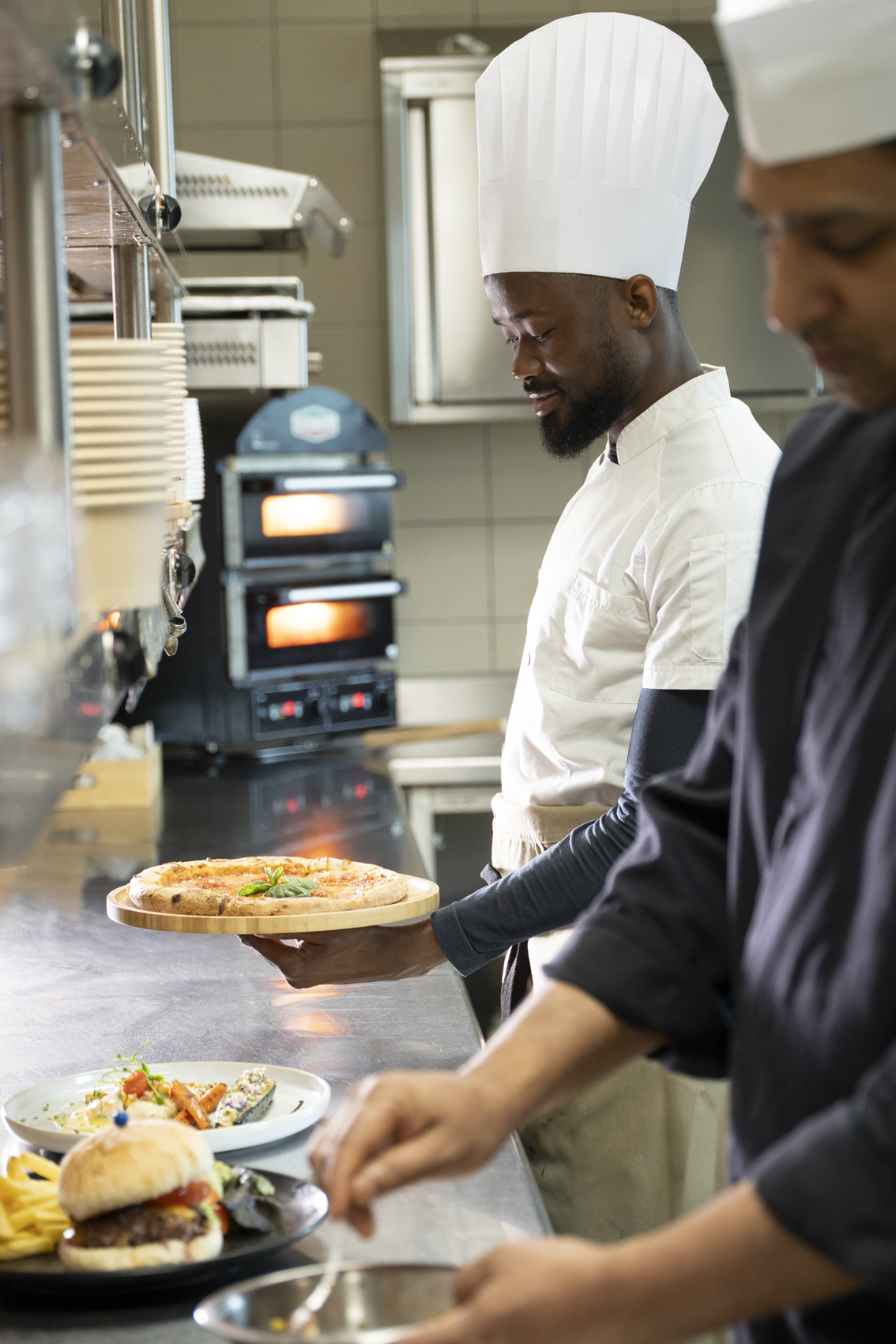 Chef holding fresh pizza in a professional kitchen with ovens and plated food.