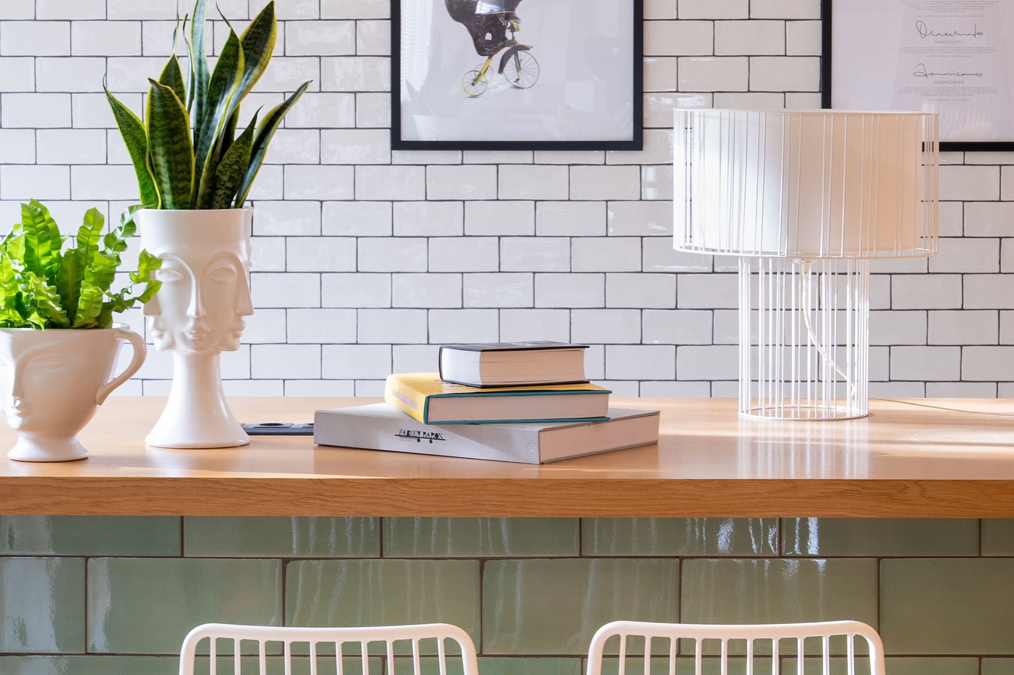 Modern cafe bar with white/green tiles, wood counter, plants, books, white stools.